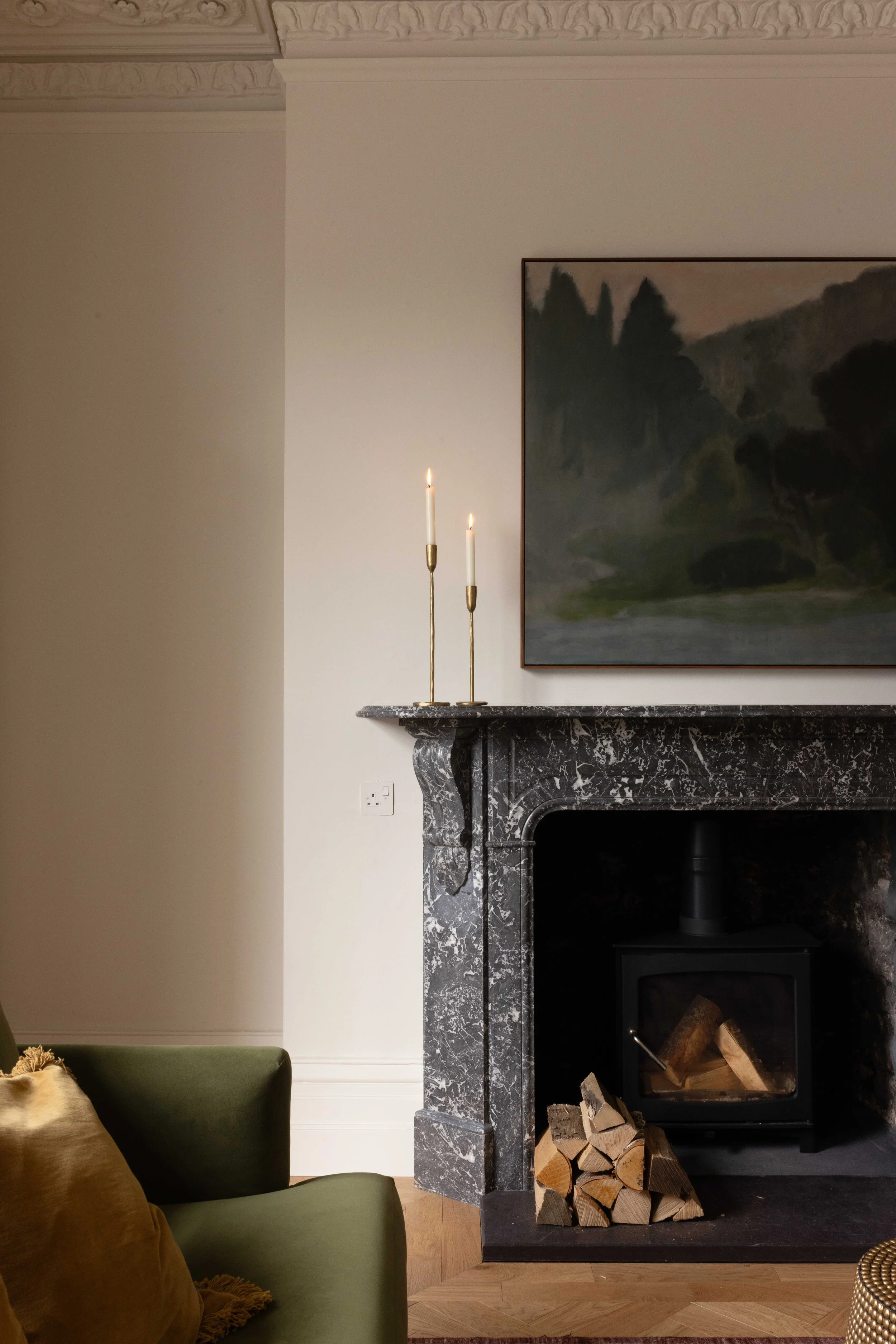 A cozy living room corner featuring a black fireplace with a pile of firewood stacked inside, a grey marble mantel, a painting of a landscape with trees hung above the mantel, and two lit candles on brass candleholders.