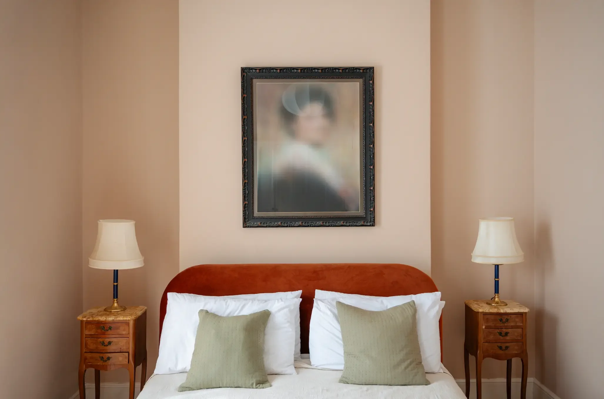 A cozy bedroom with a bed, two wooden nightstands with lamps, five pillows on the bed, a framed mirror on the wall, and beige walls.