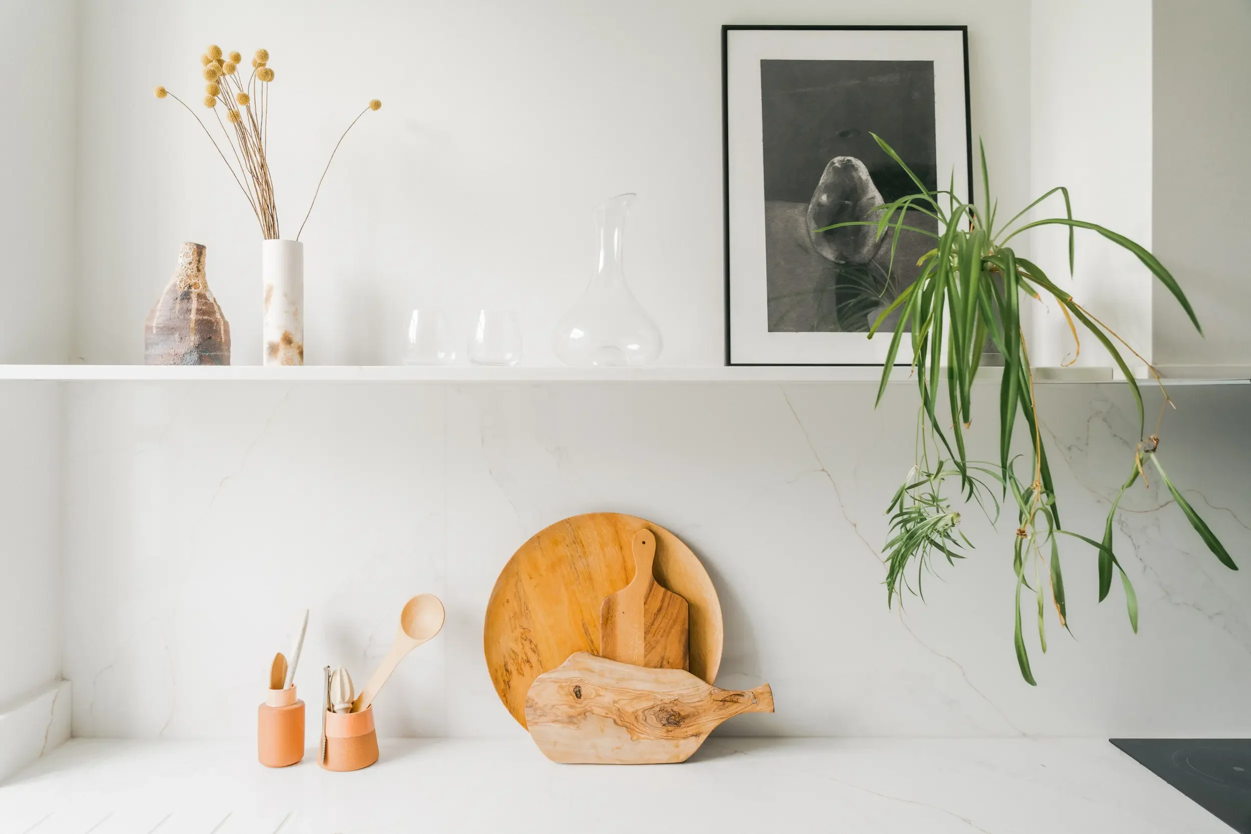 Minimalist kitchen counter with wooden cutting boards, utensils, a potted plant, framed black and white photo, and decorative vases with dried flowers.