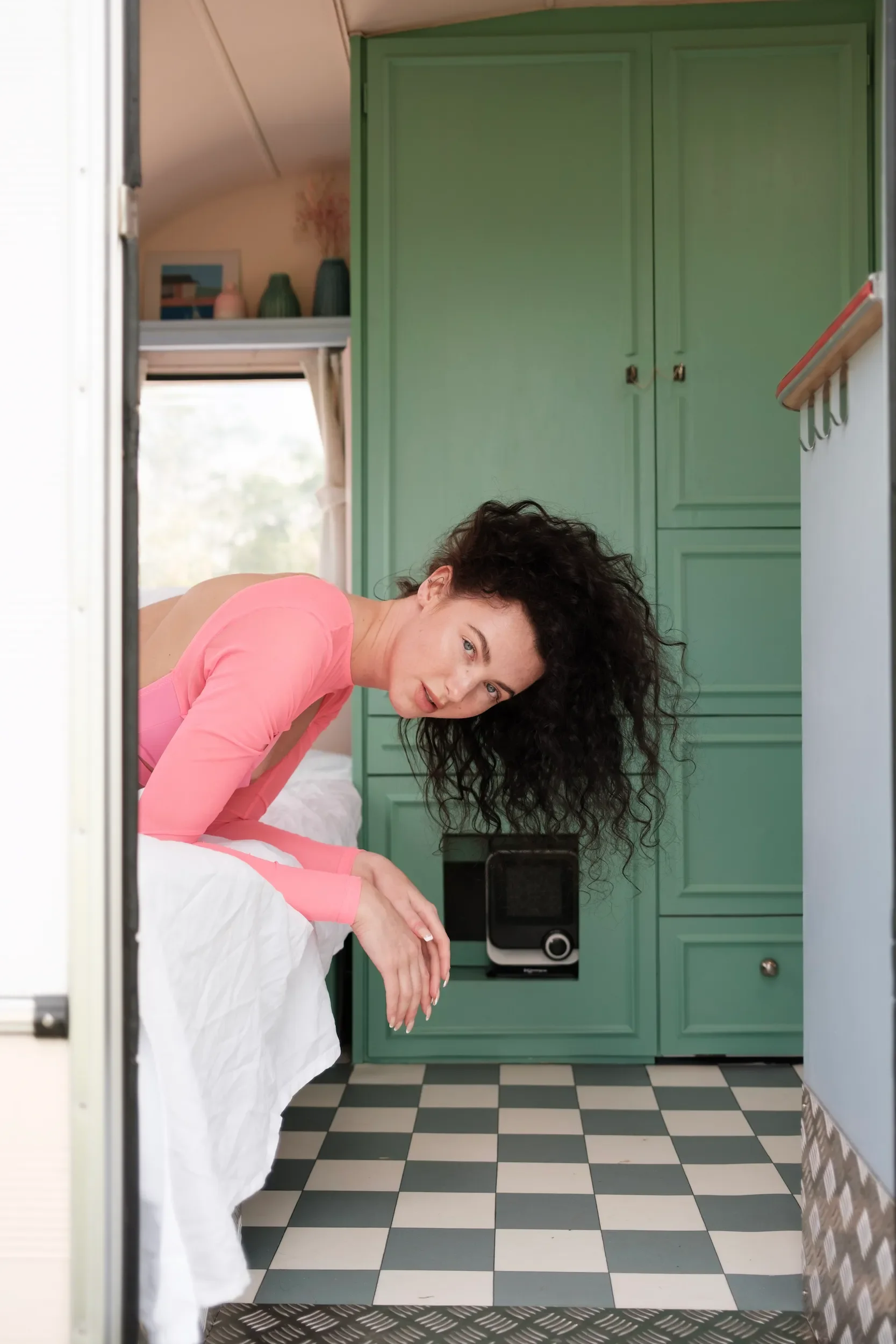 A woman with curly dark hair and a pink shirt sitting on a bed in front of a mirror, looking at her reflection, in a room with green cabinets and a checkered black and white floor.