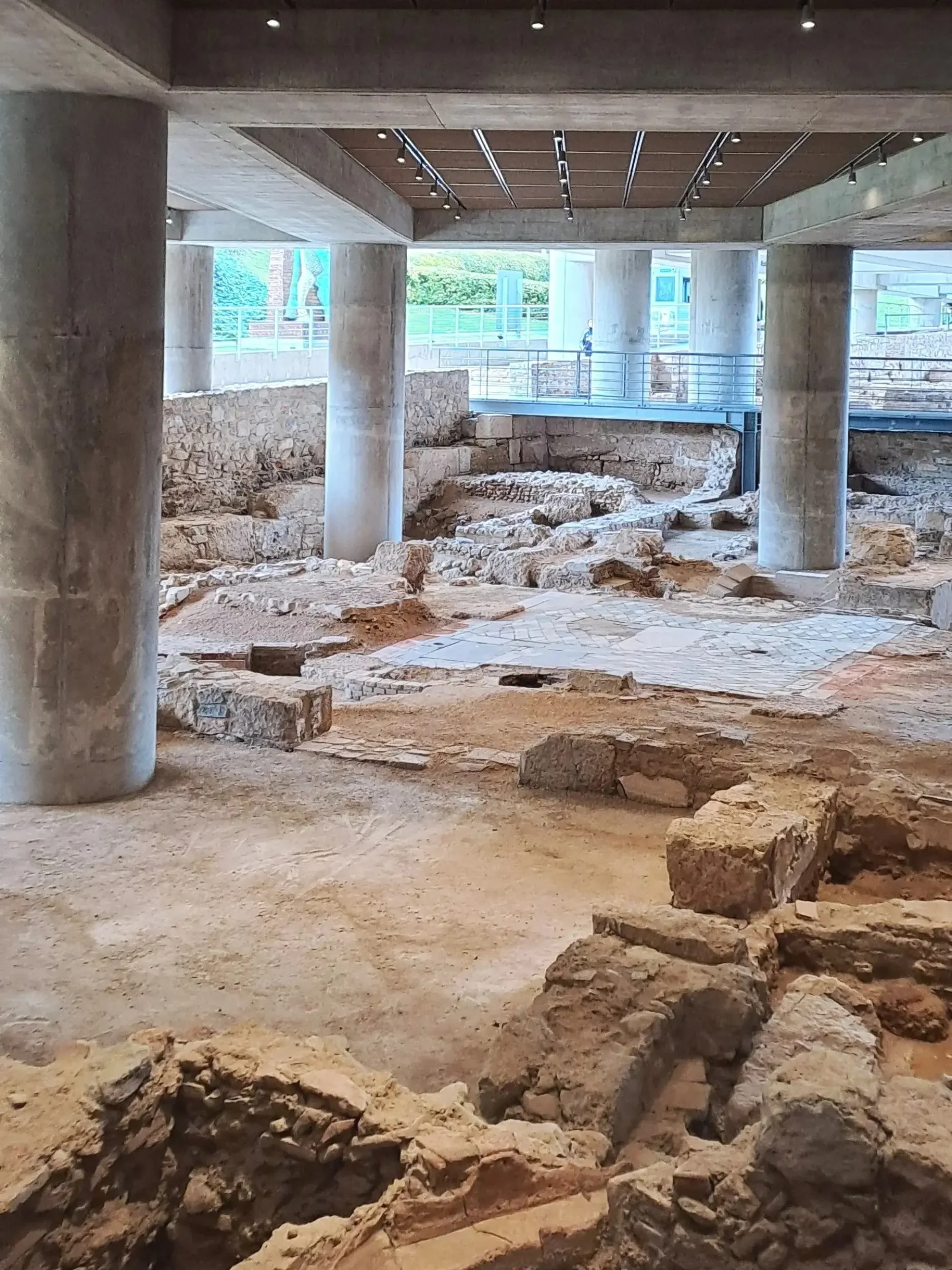 Archaeological site with stone ruins and modern concrete infrastructure, under a bridge or overpass.