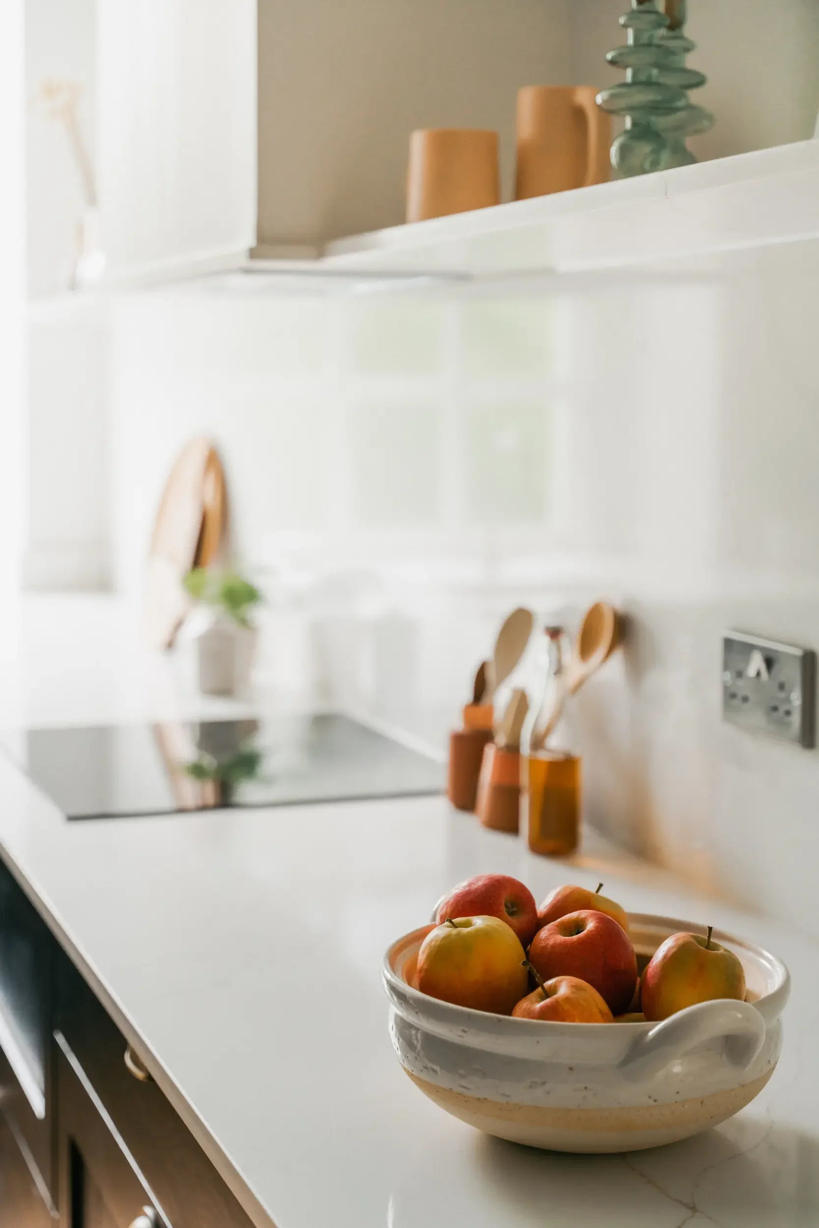 A bowl of apples on a kitchen countertop with utensils in holders, a potted plant, and decorative items on a shelf in the background.