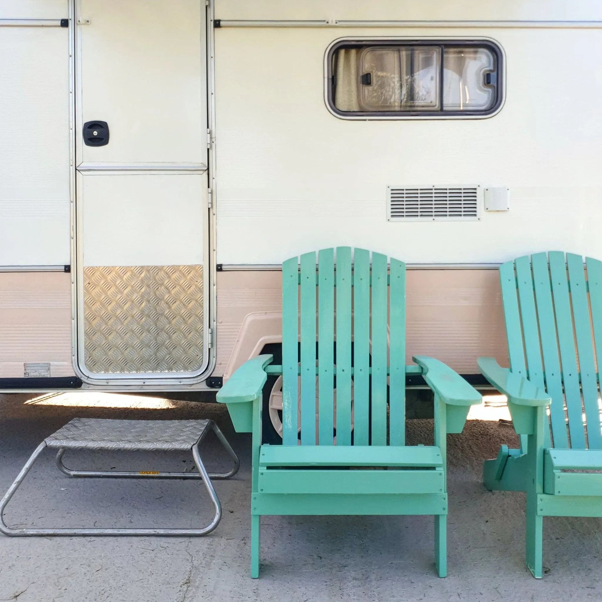 Outdoor scene featuring a pale blue Adirondack chair in front of a light-colored trailer with a window, a small garage-style door, and metal trim, on a gravel surface with another chair and a small metal platform nearby.