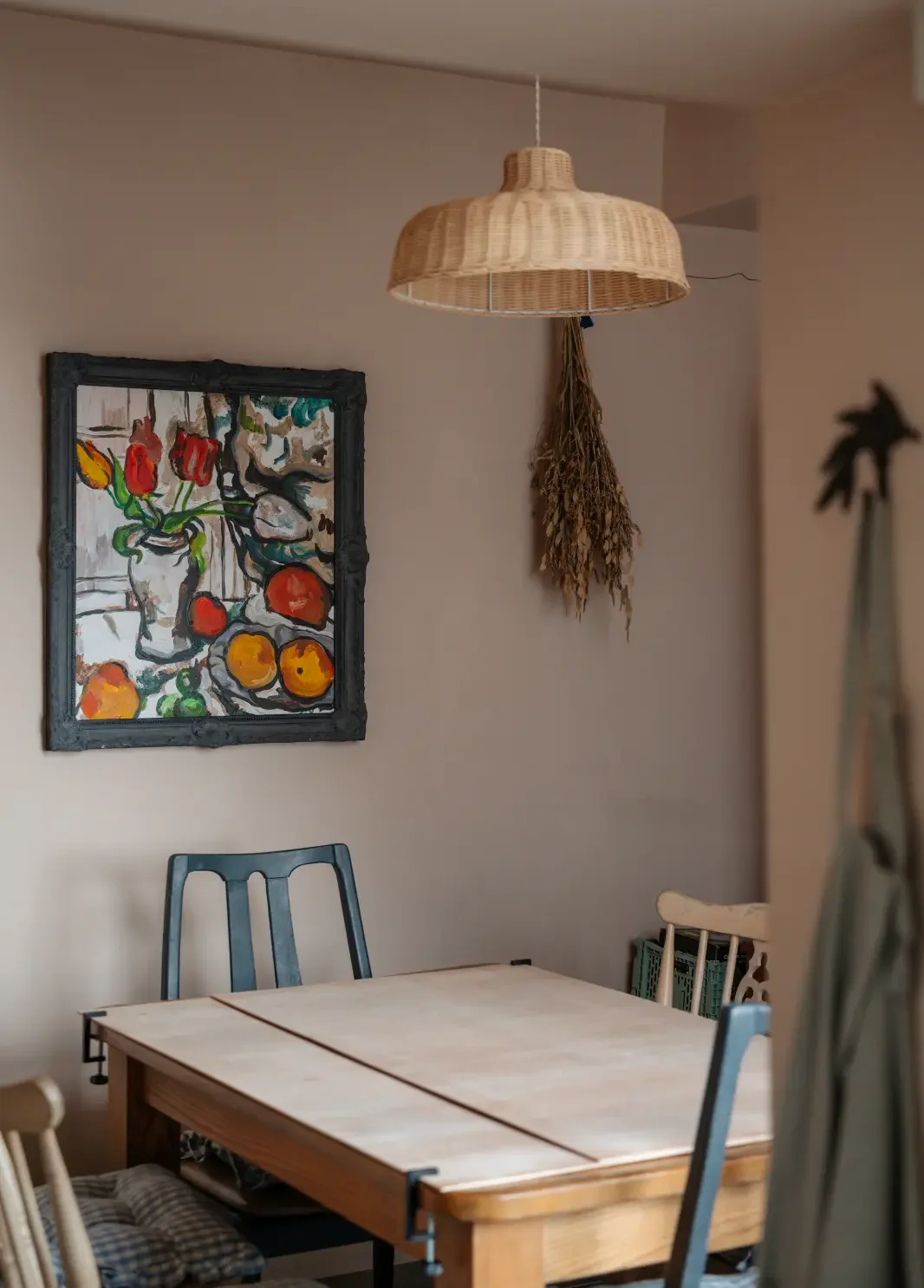 A dining room with a wooden table and mixed chairs. A hanging woven lampshade suspended above the table. A colorful still life painting of fruits and flowers on the wall, and dried herbs hanging from the ceiling. Part of a mirror and a plant in the foreground.