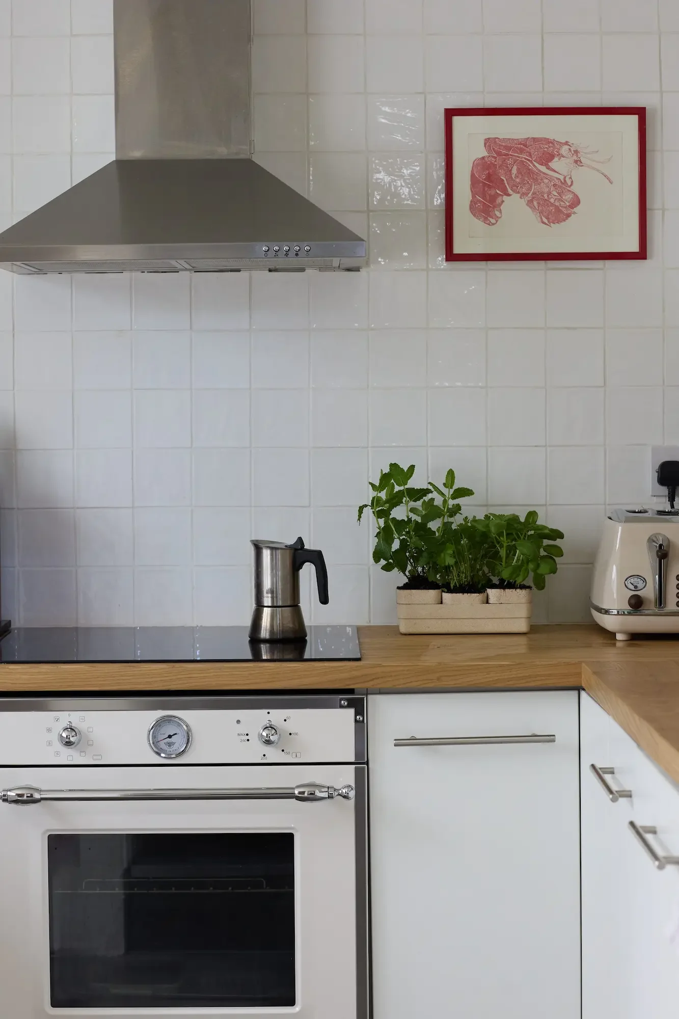 Kitchen with white tiled wall, stainless steel range hood, electric stove, white cabinetry, a potted herb plant on wooden countertop, a framed illustration of a lobster on the wall, a kettle, and a toaster.