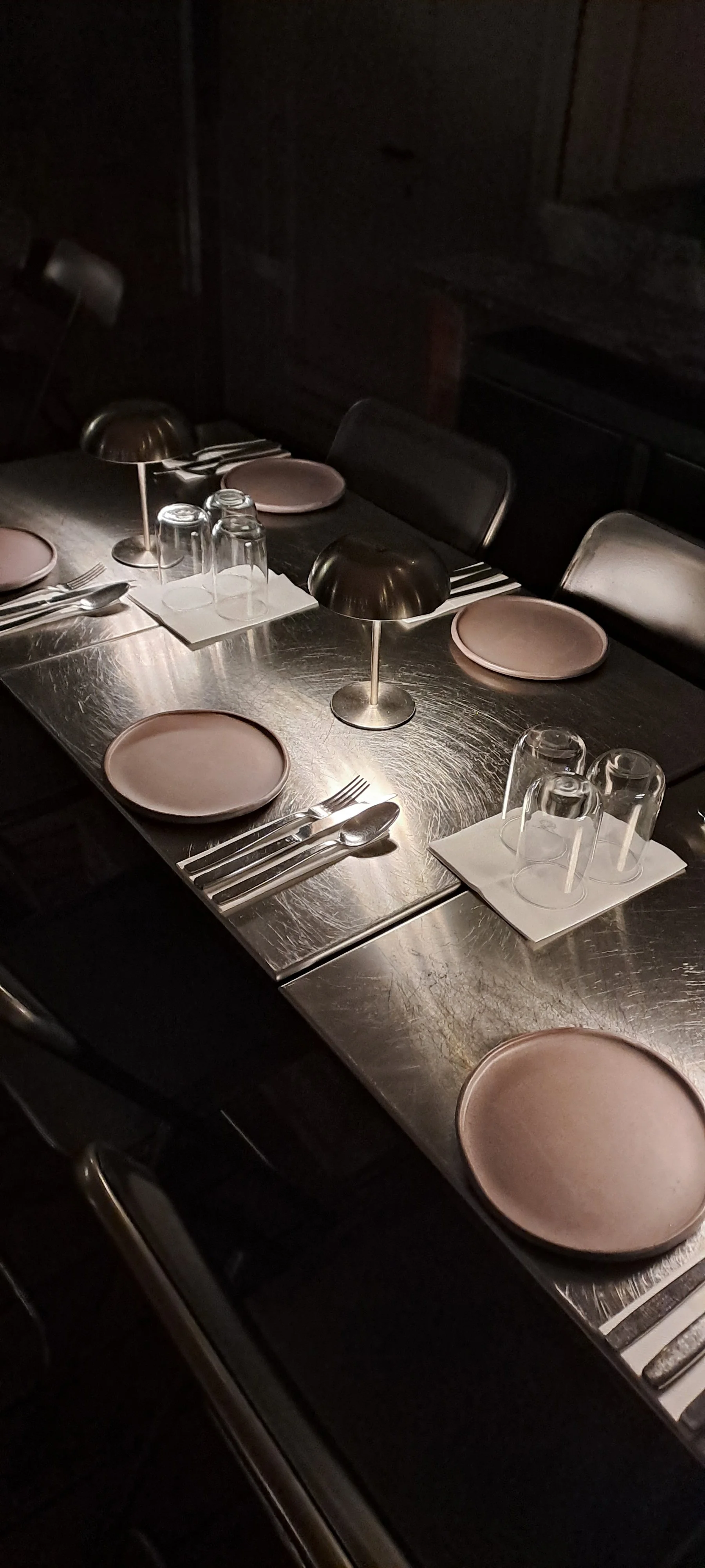 A dimly lit restaurant table set with pink plates, stainless steel utensils, upside-down drinking glasses on white napkins, and metallic table lamps.