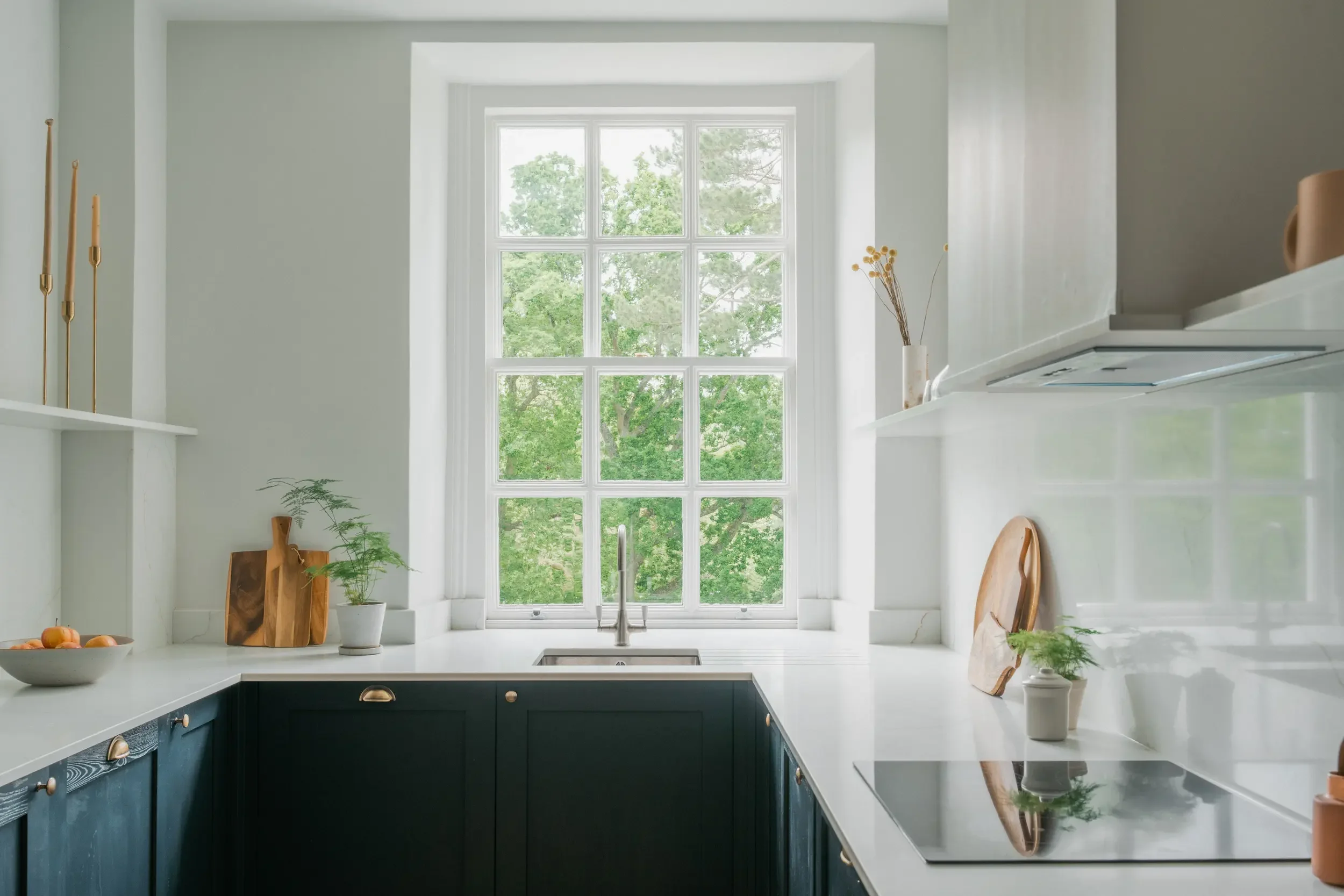Modern kitchen with large window overlooking green trees, white countertops, black lower cabinets, and minimal decor including plants, boards, and a bowl of fruit.
