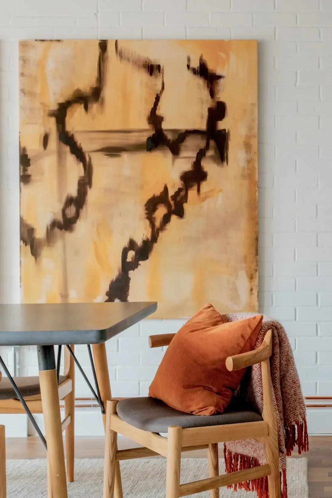A modern dining room with a white brick wall, a wooden chair with a rust-colored pillow, a table, and a large abstract painting on the wall.