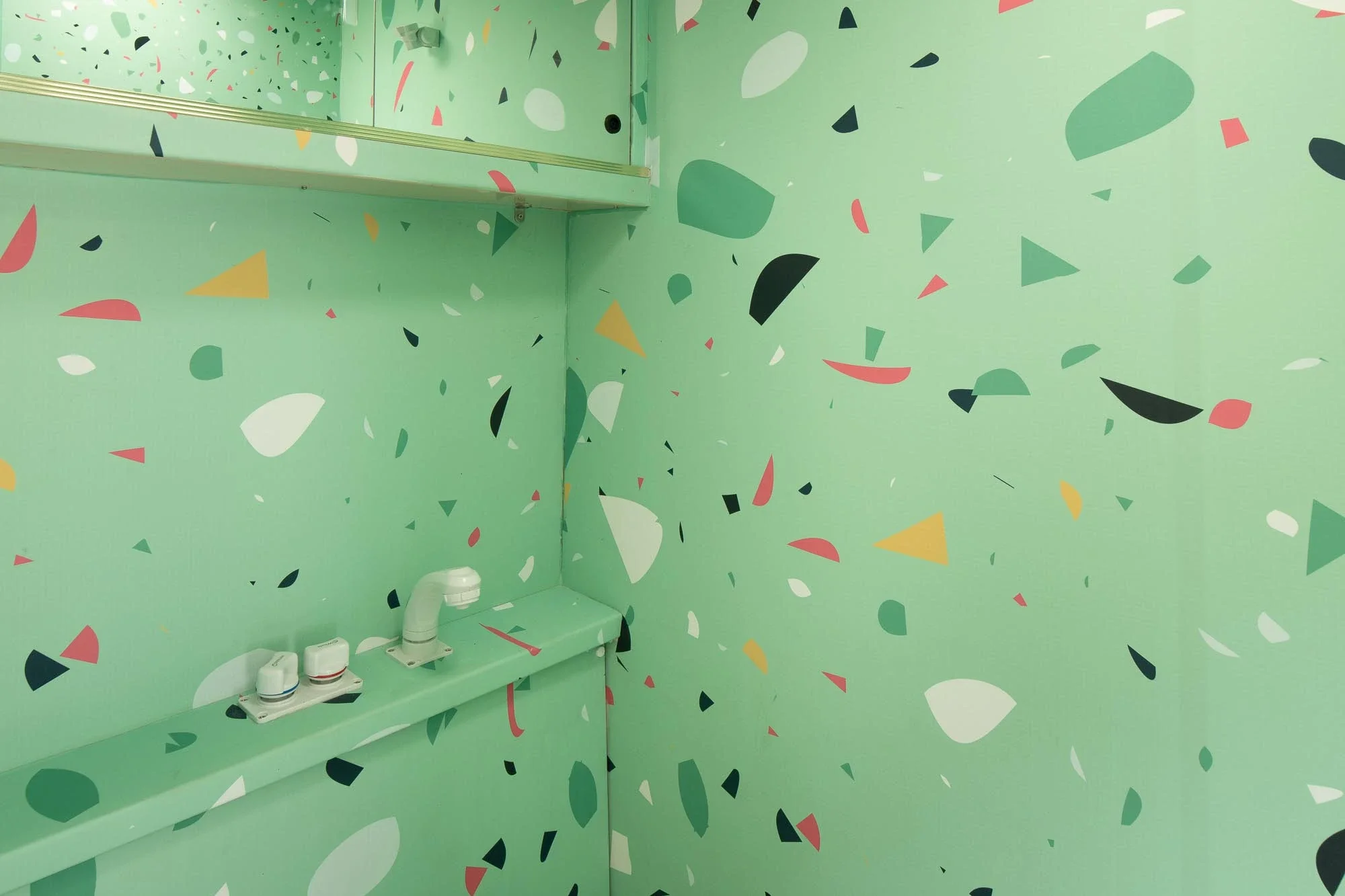 A green room decorated with multicolored confetti patterns, featuring a mirror, a small sink, and a faucet.