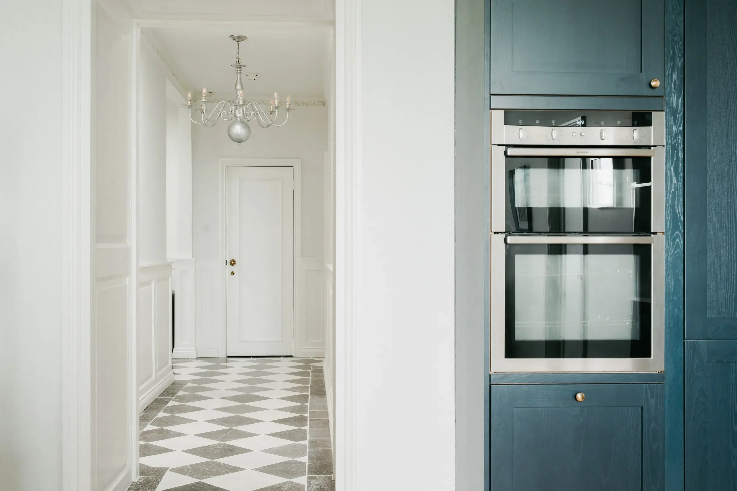 Interior view of a house showing a kitchen and a hallway with a chandelier. The kitchen has built-in stainless steel oven and blue cabinets. The hallway has tiled checkered flooring and white walls with wainscoting.