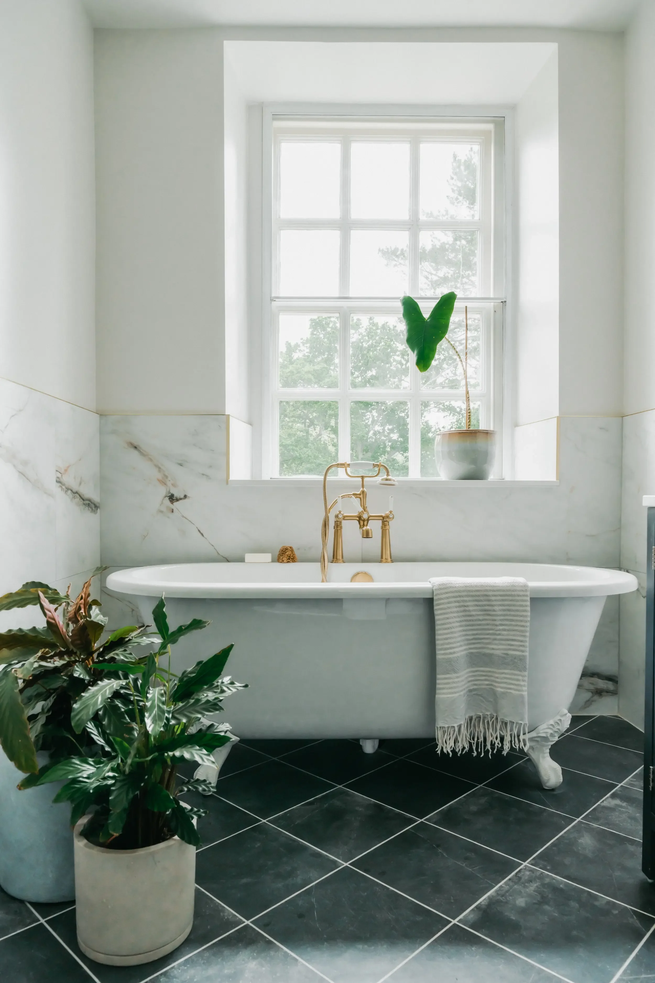 A clean, modern bathroom with a vintage clawfoot bathtub in front of a large window, decorated with green plants and a white towel hanging on the side.