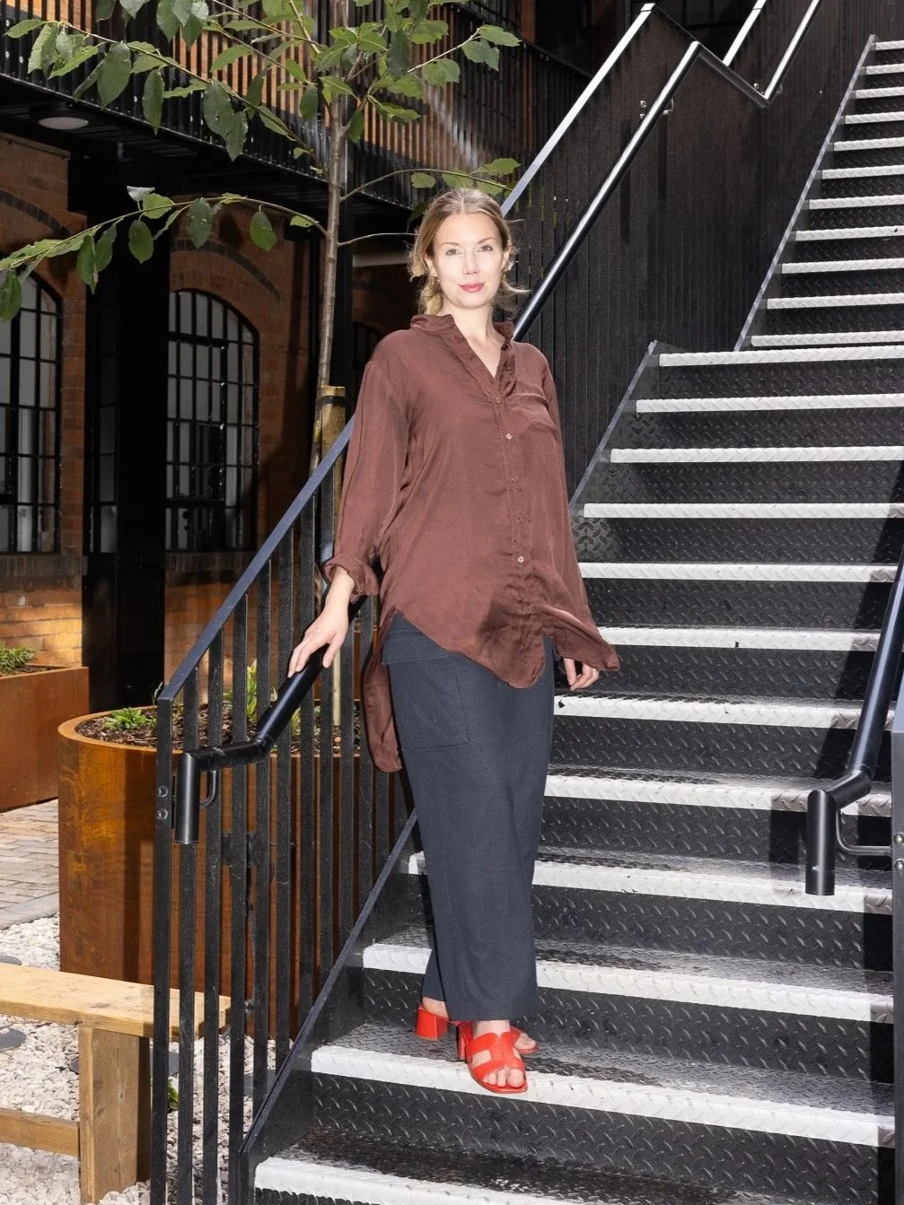 A woman standing on an outdoor black metal staircase, holding the railing with her left hand. She is wearing a loose brown button-up shirt, dark wide-leg pants, and red heeled sandals. Behind her is a tree with green leaves and a building with brick walls and large black-framed windows.