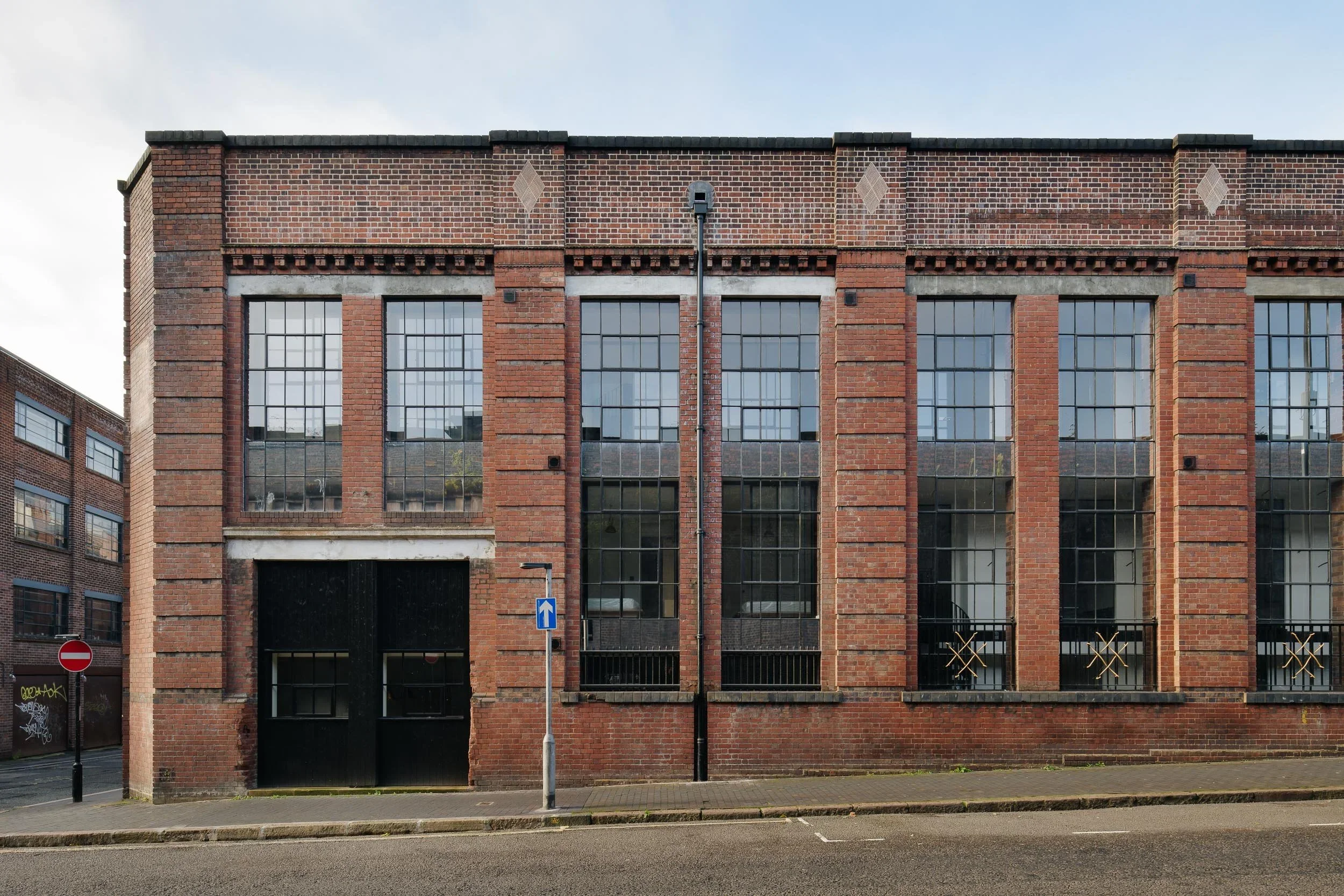 A red brick building with large windows and decorative brickwork, a street with a no-entry sign, and a tall streetlamp in the foreground.