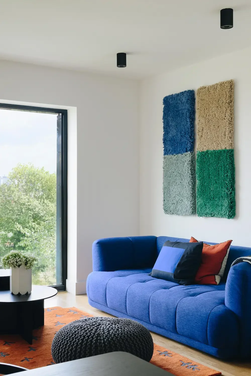 Living room with blue sofa, colorful cushions, black round ottoman, orange rug, potted plant, and abstract wall art, next to large window showing greenery outside.