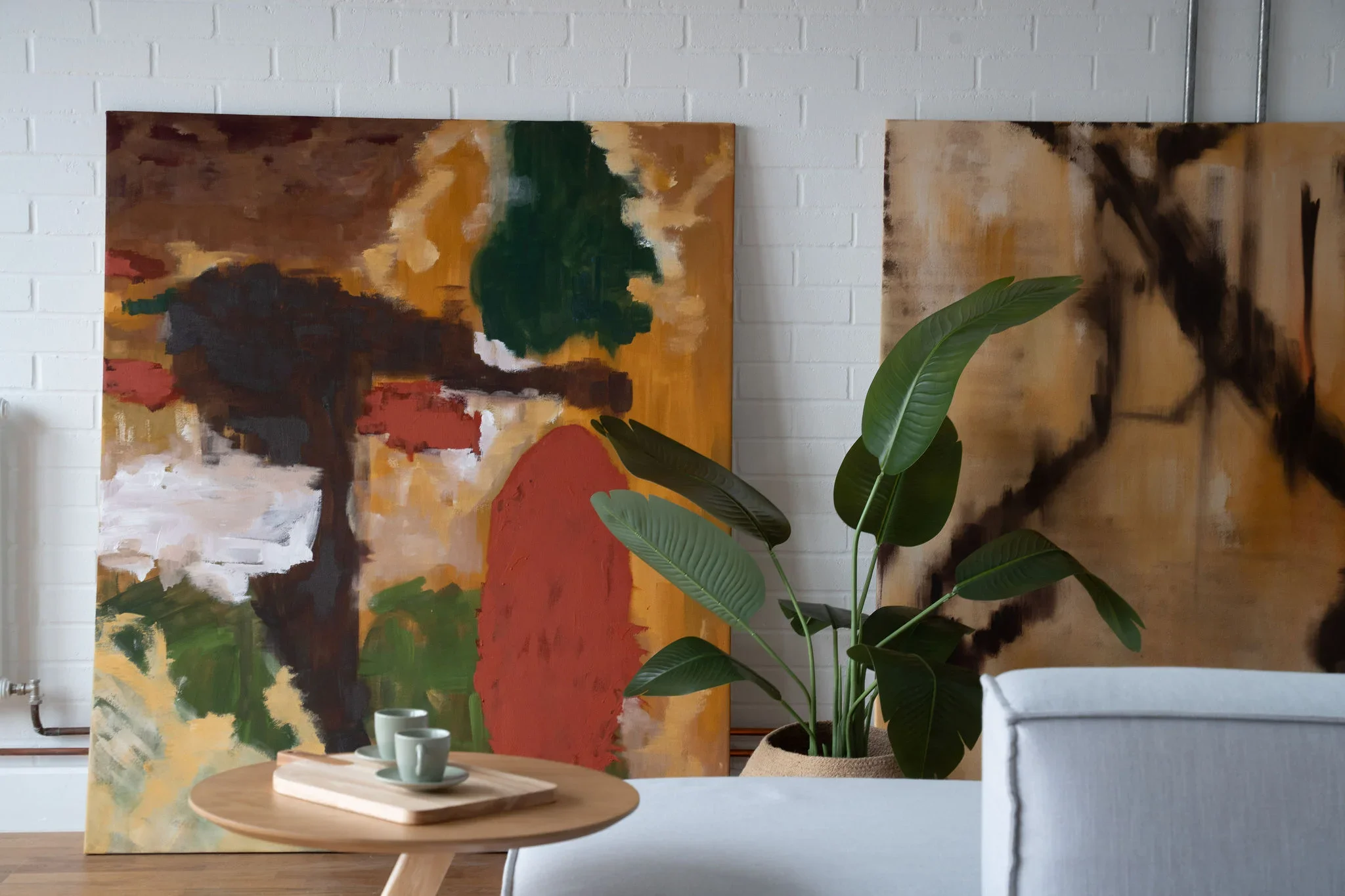 Interior of an art studio or gallery with abstract paintings on a white brick wall, a green potted plant, and a round wooden table with small green cups.