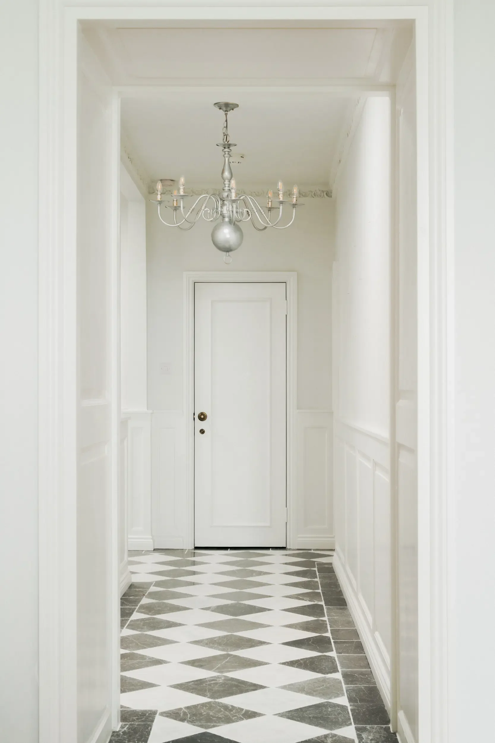 A narrow, white hallway with wainscoting on the walls, a diamond-patterned black and white marble floor, a white door at the end, and an ornate chandelier hanging from the ceiling.