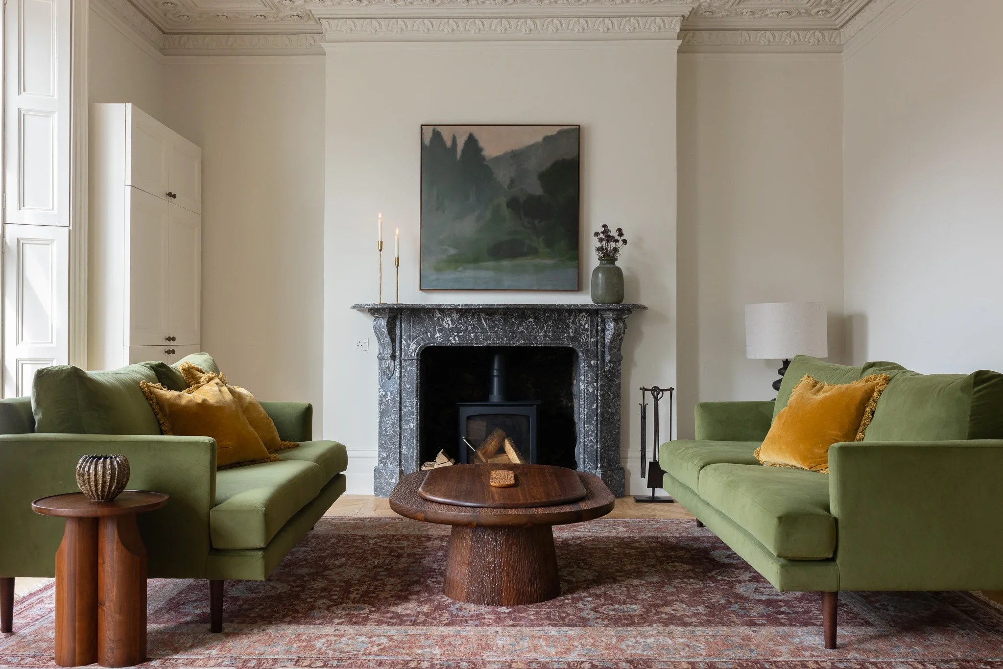 A living room with two green sofas with throw pillows facing each other, a wooden coffee table in the center, a dark marble fireplace with a painting above, decorated with candles and a vase on the mantle, a lamp on the side, and a patterned rug on the floor.