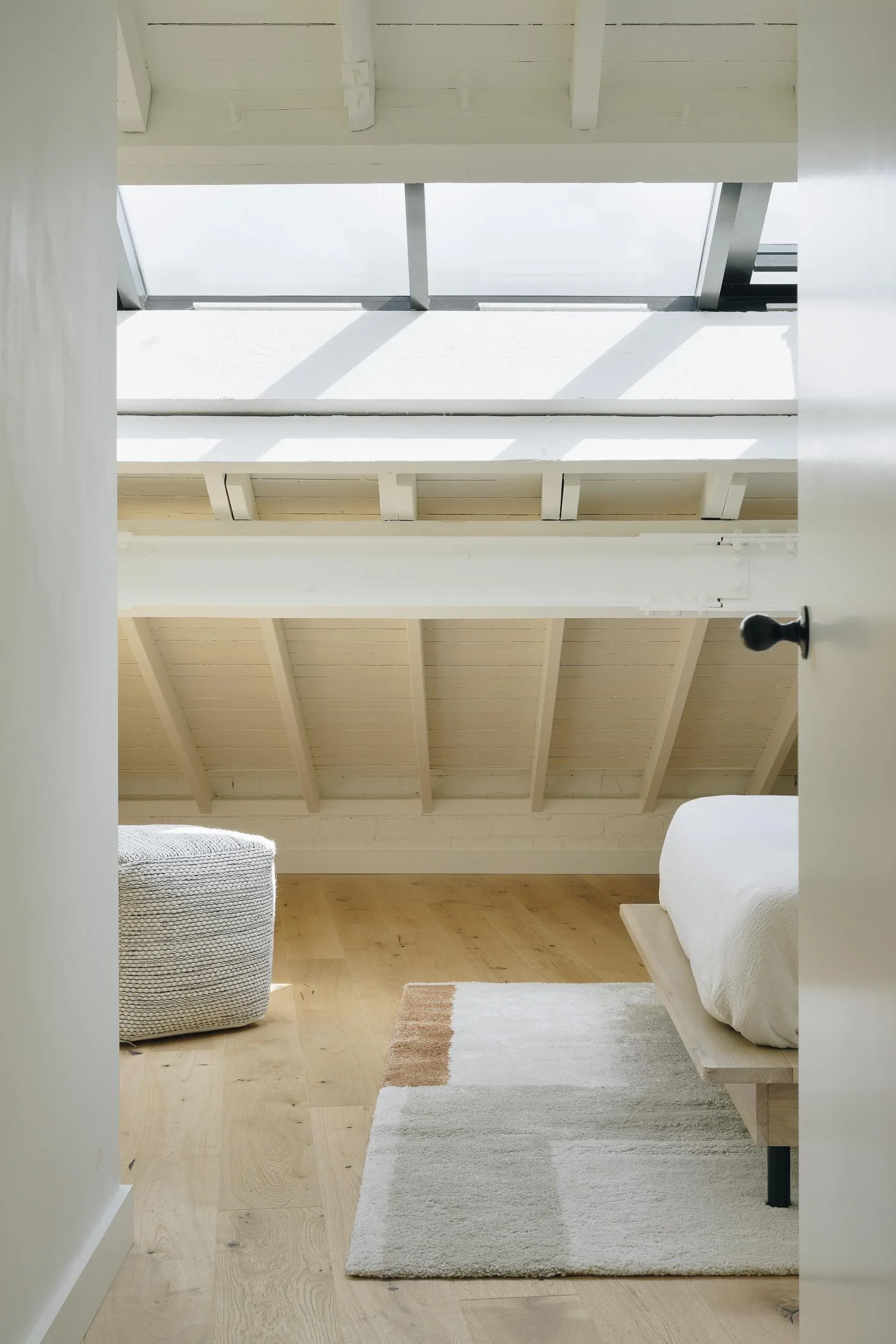 A minimalist bedroom with a slanted white ceiling, wooden floor, and simple furniture. There is a woven ottoman and a bed with a curved white blanket, a small wooden side table, and a light-colored rug.