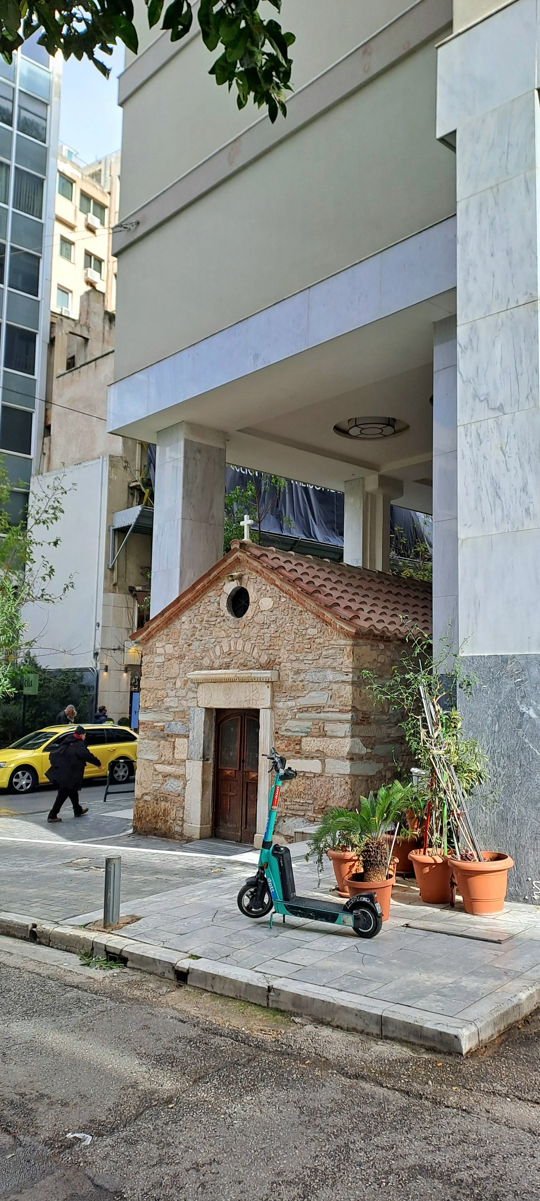 Small stone chapel with wooden door, red tile roof, and a small window at the front, surrounded by potted plants, scooter, and city street with modern buildings and a yellow car in the background.