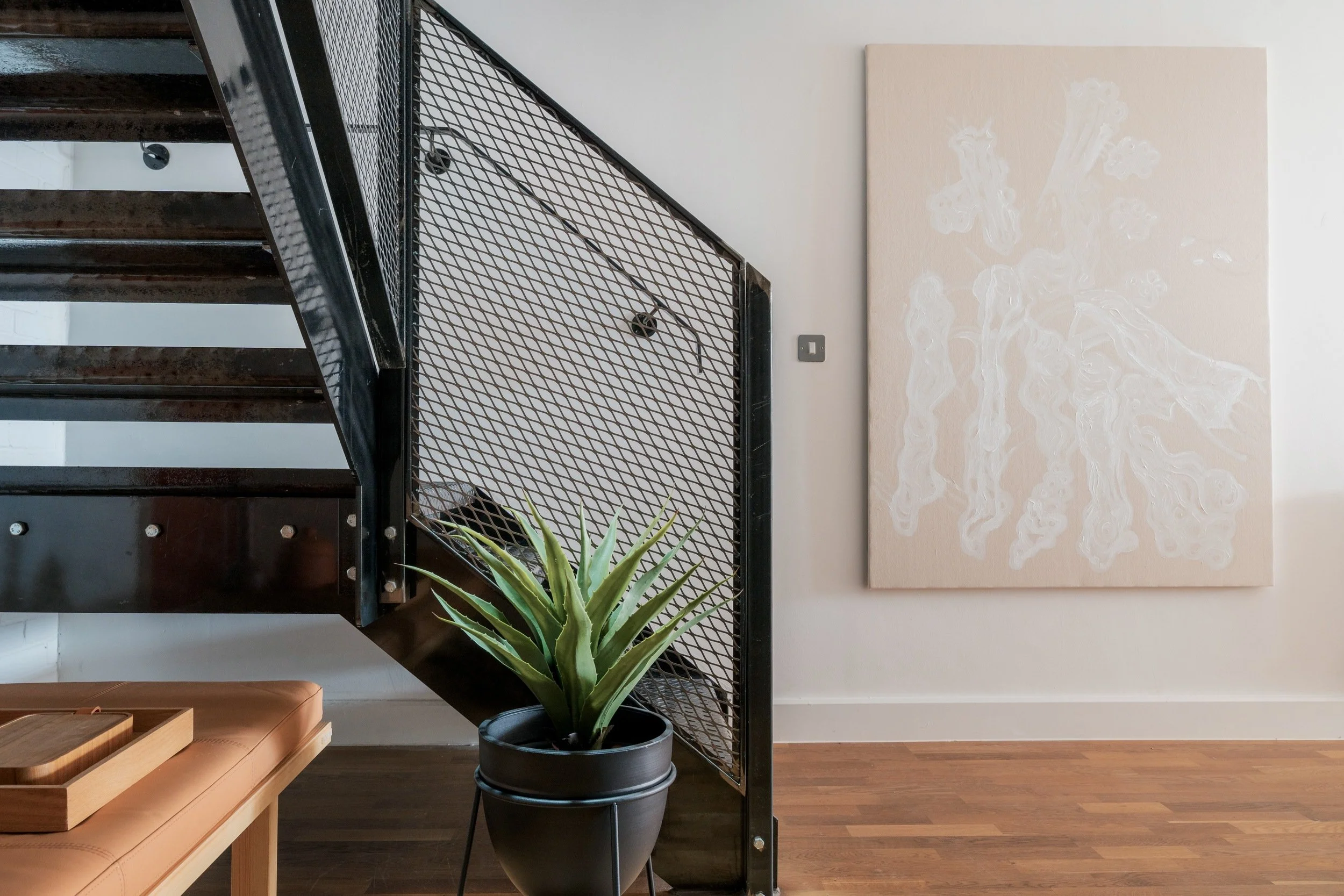 Industrial black metal staircase. Neutral large formal canvas on a white wall. Green plant in a large format pot. Artwork by Aly Grimes, founder and director of San Stae Interior Design and Art Studio.