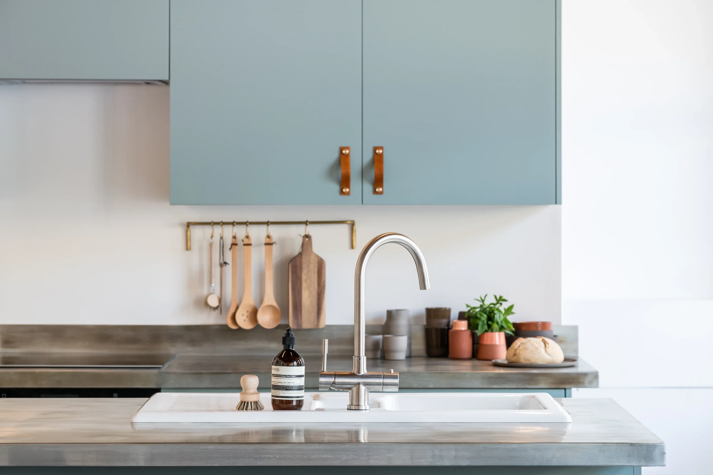 Modern kitchen with light blue cabinets, wooden handles, hanging utensils, a sink, and various kitchen tools and plants on the countertop.