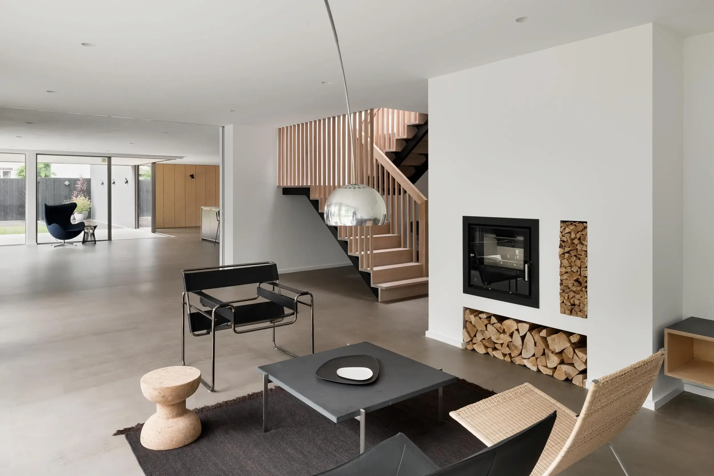 Modern living room with a staircase featuring wooden railing, a fireplace with stacked wood storage, a black chair and a black coffee table on a dark rug, and large sliding glass doors leading outside to a green yard.