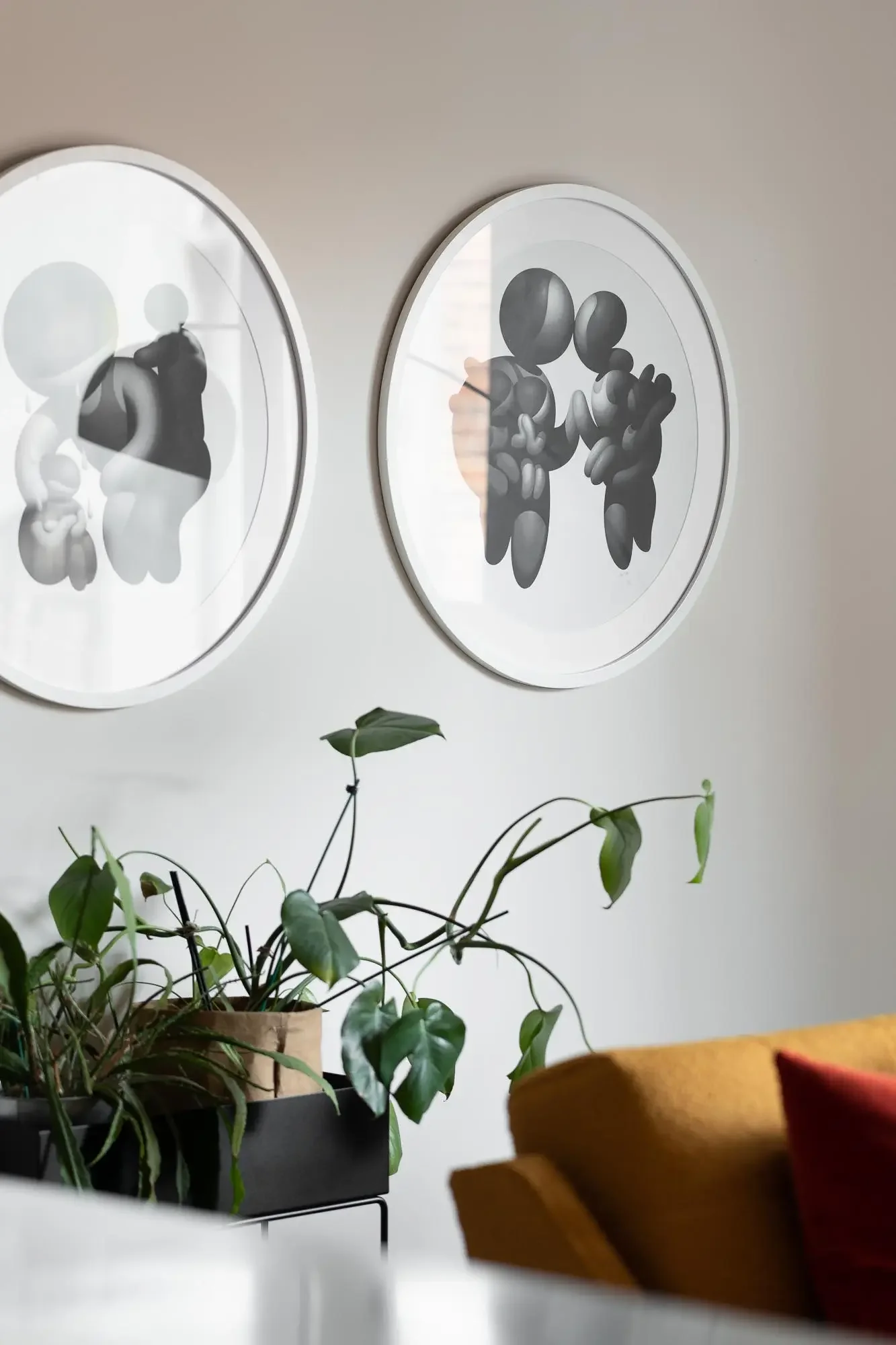 Indoor scene with a potted plant on a black stand, two framed black-and-white art pieces on the wall, and part of a wooden chair or furniture in the foreground.