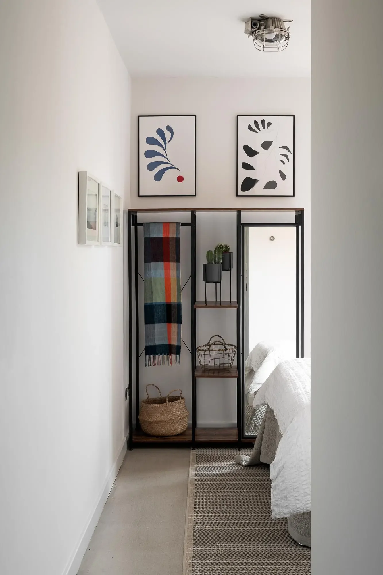 A bedroom hallway with white walls, decorated with framed artwork. There is a black metal and wood shelving unit holding a multicolored plaid blanket, small cacti in black pots, and a basket. The bed with a white comforter is visible on the right side.