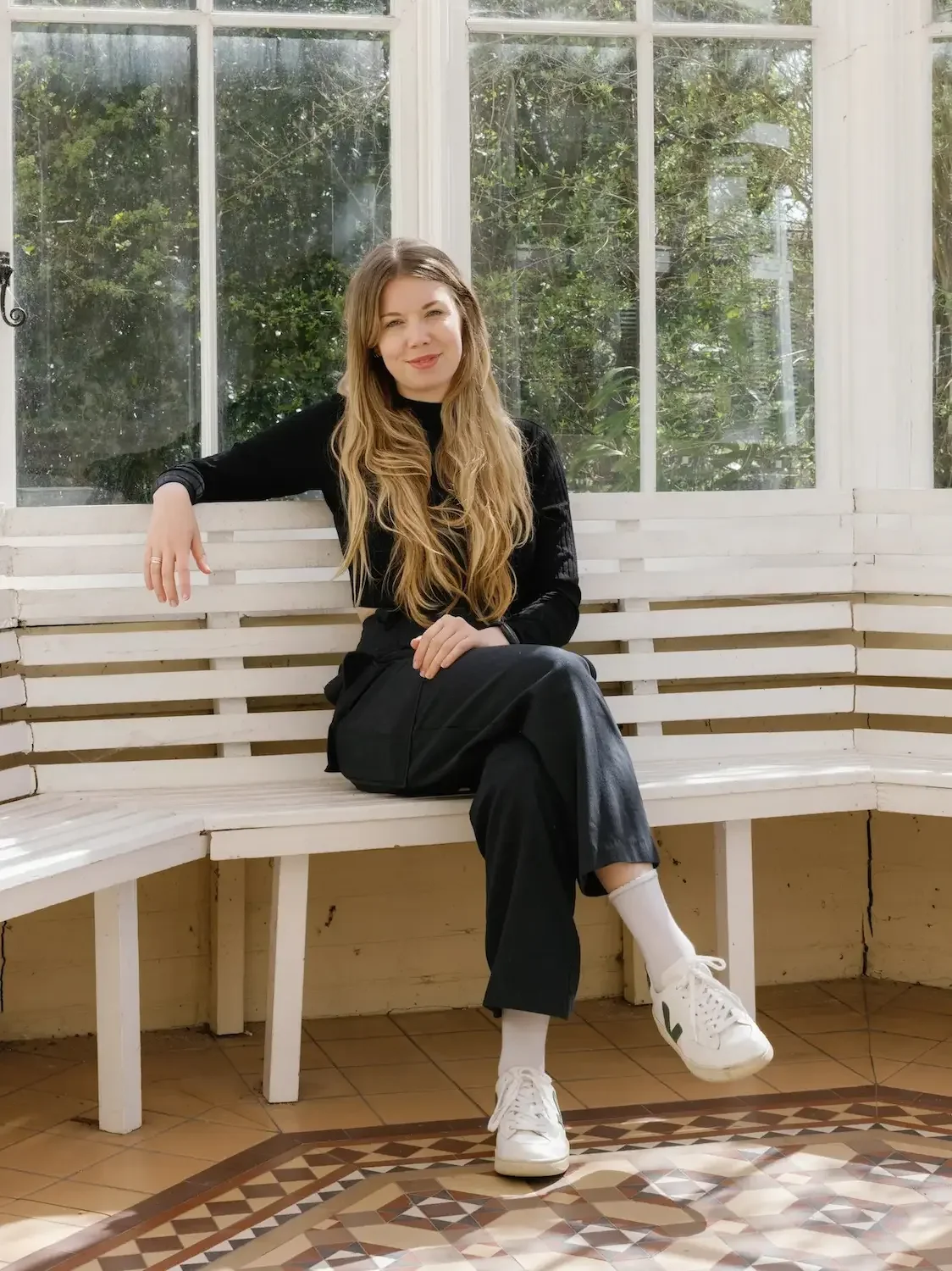 Young woman sitting on a white bench in front of large windows with greenery outside, wearing black outfit and white sneakers, smiling and winking.