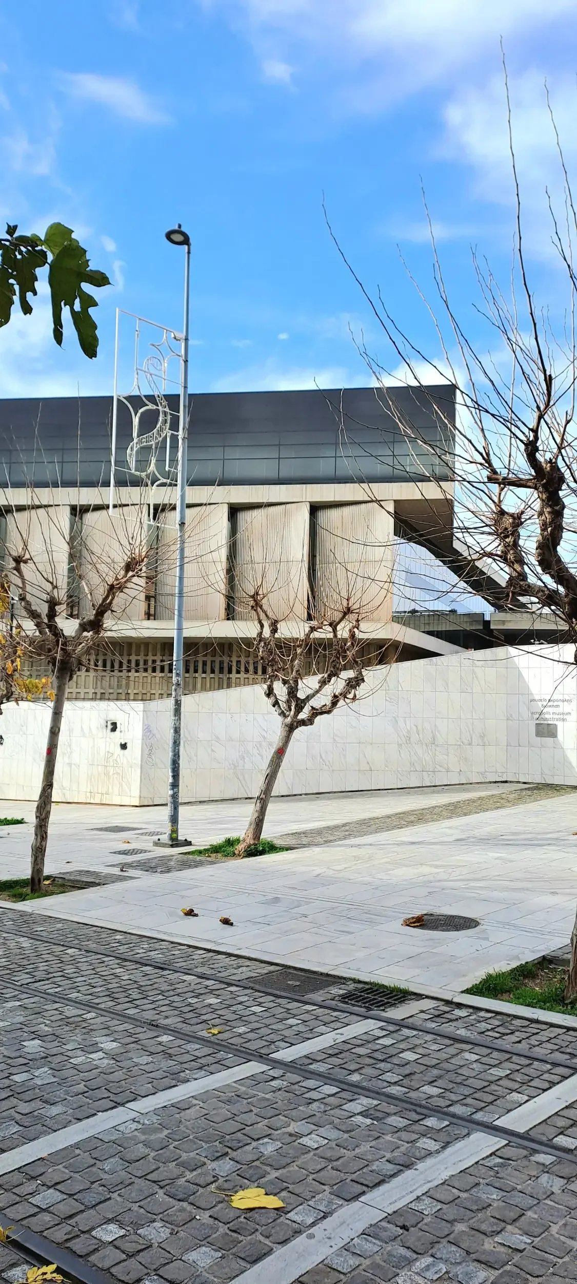 A modern building with wood paneling, surrounded by leafless trees and a paved walkway, under a blue sky with some clouds.