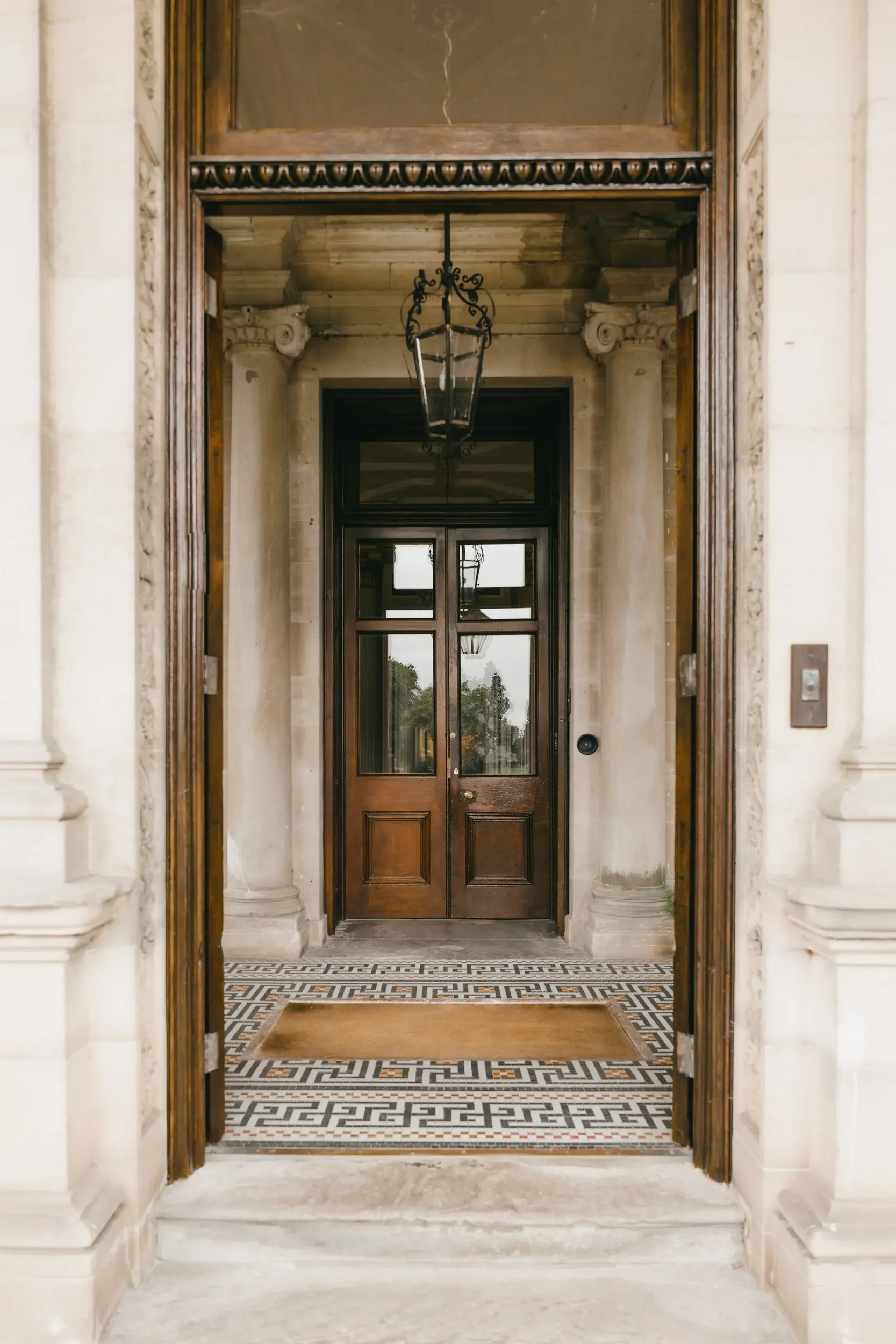 Wooden front door with glass panels, framed by columns, under a decorative lintel, with a patterned tile floor and a lantern hanging overhead.