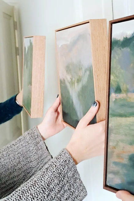 People holding framed landscape paintings with mountain and greenery scenes.