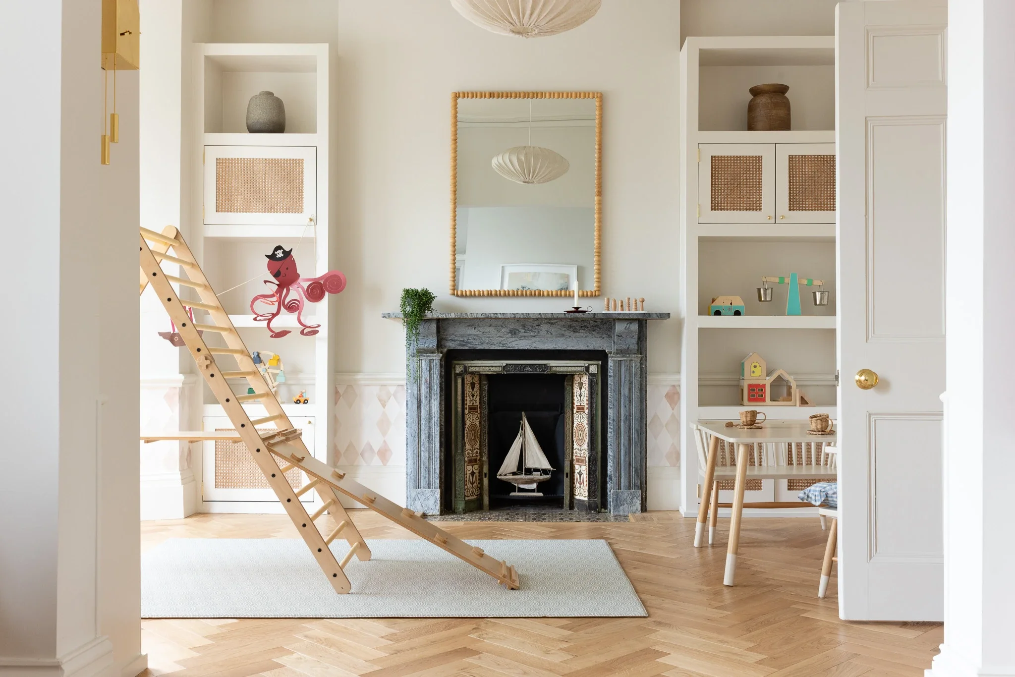 A cozy living room with a marble fireplace, shelves on either side, a large mirror above the fireplace, and a wooden slide toy with a pink octopus hanging decorations in front. There is a small table with chairs on the right and a beige rug on a herringbone wooden floor.