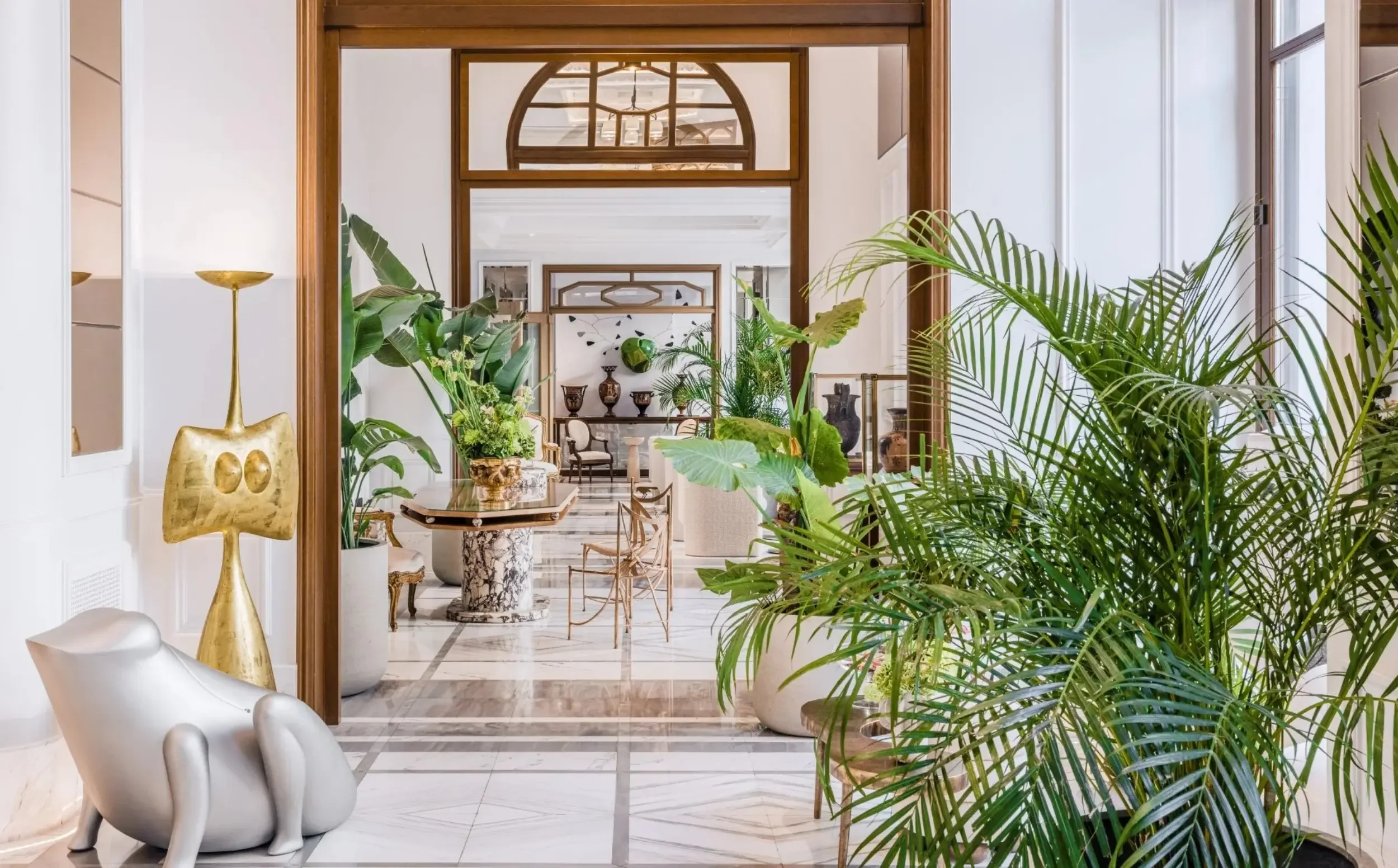 Elegant interior of a building with large windows, indoor plants, and artistic decor including a silver sculpture and a gold abstract lamp.