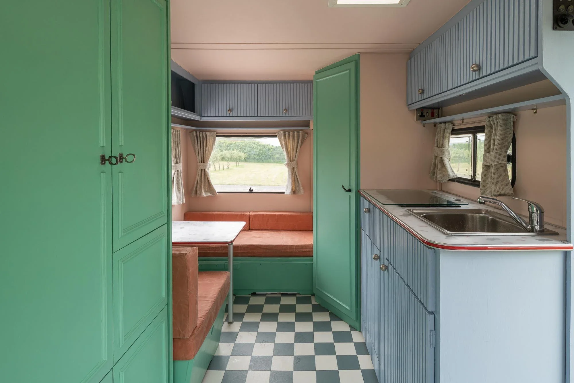 Interior of a vintage camper with green cabinets, a small kitchen area with a stainless steel sink, blue and white striped cabinet doors, a checkered floor, and a sitting area with a window, curtains, and a built-in bench with orange cushions.