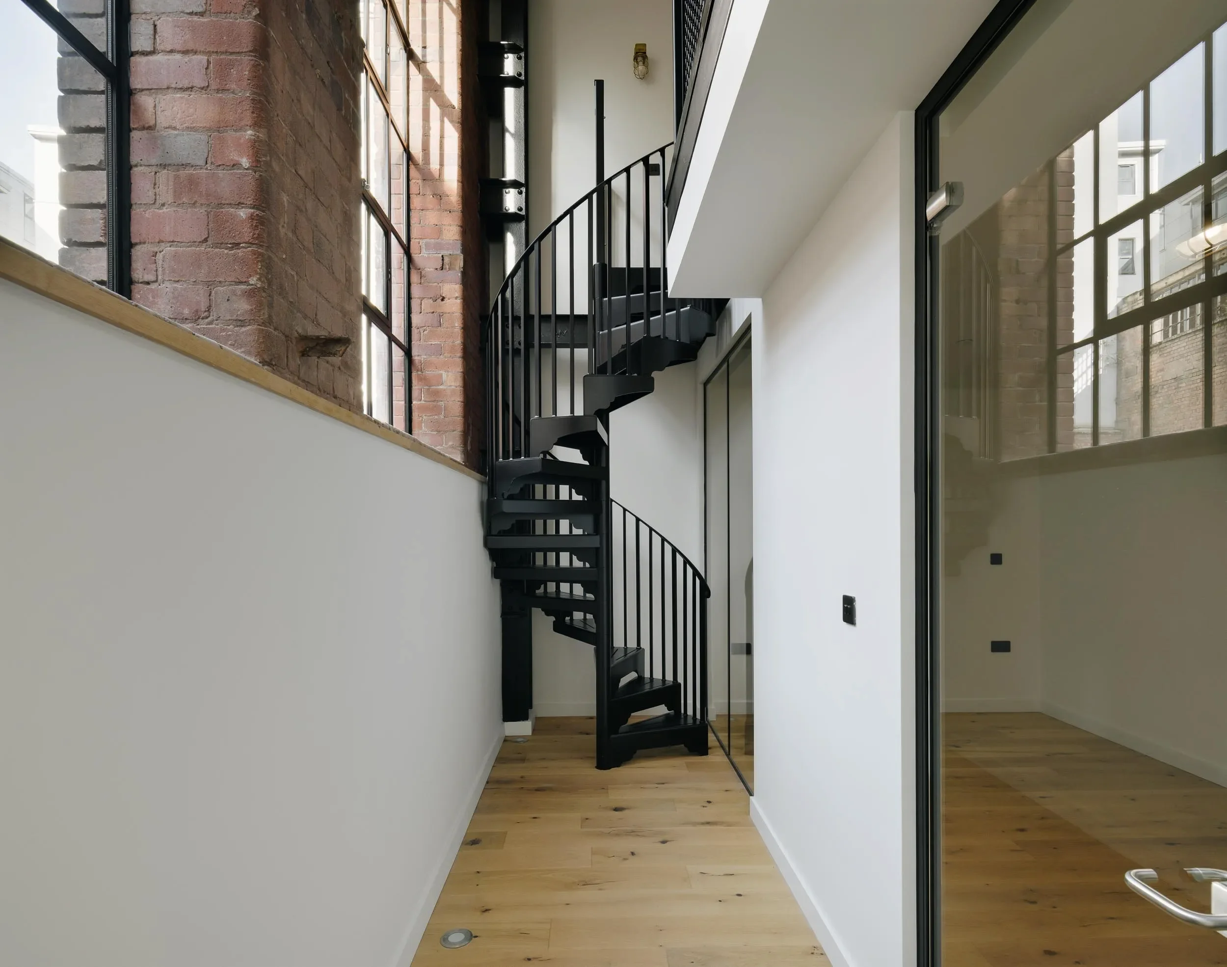 Interior view of a modern building featuring a black metal spiral staircase, exposed brick wall, large windows with black frames, and wooden flooring.
