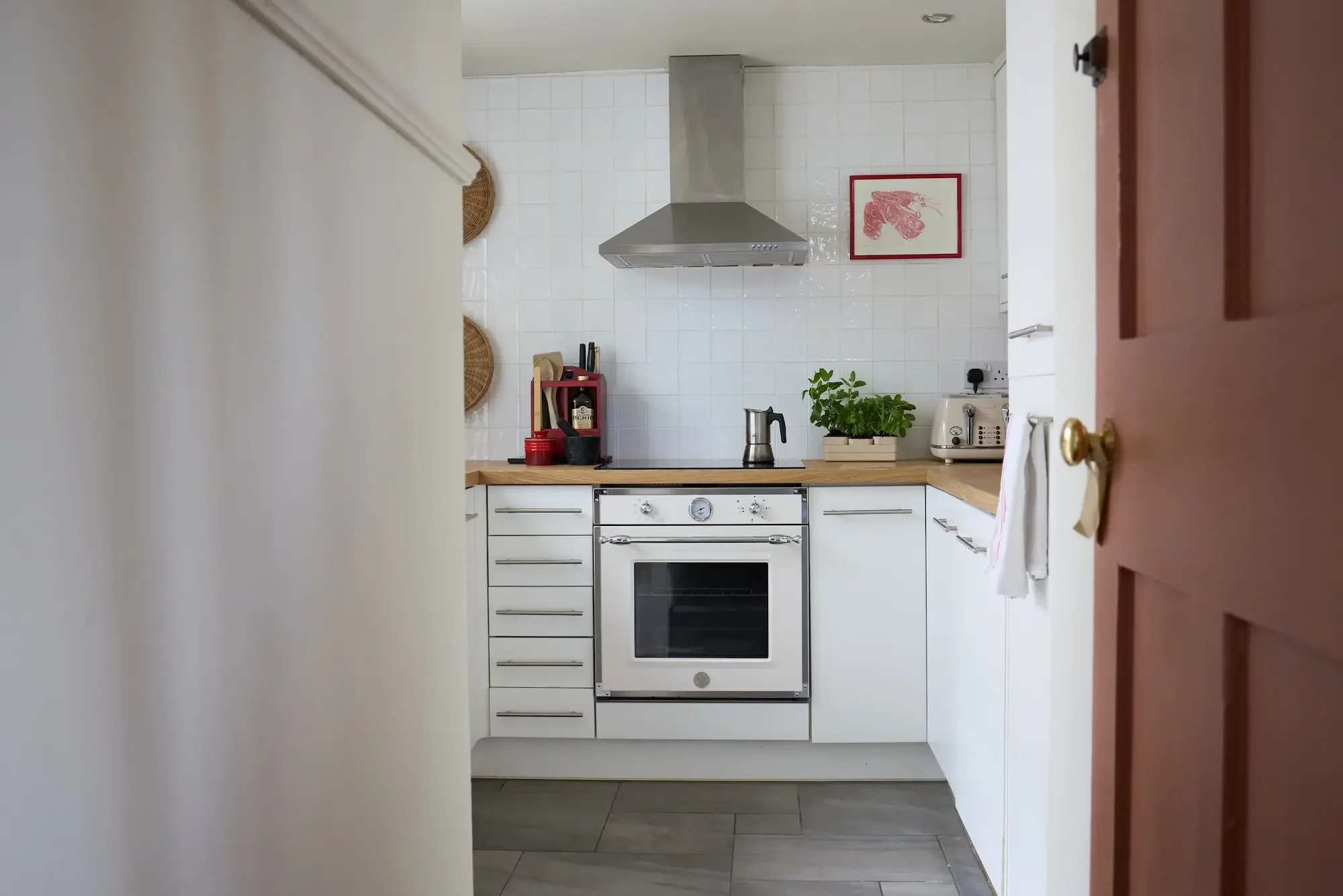 View of a modern kitchen with a white oven, stovetop, and stainless steel range hood. The kitchen has white cabinets, a wooden countertop, and a white tiled backsplash. On the countertop, there is a potted plant, a toaster, and a red shelf with bottles and kitchen utensils. A framed picture of a lobster hangs on the wall above the countertop. The image is taken from partly behind a door, with the door frame visible on the right side.