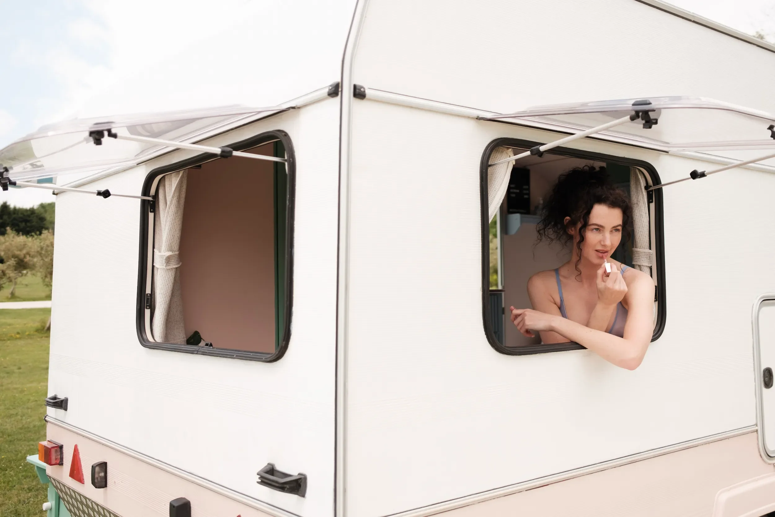 A woman leaning out of a camper window, looking at something reflectively while holding her chin, with a grassy outdoor setting in the background.