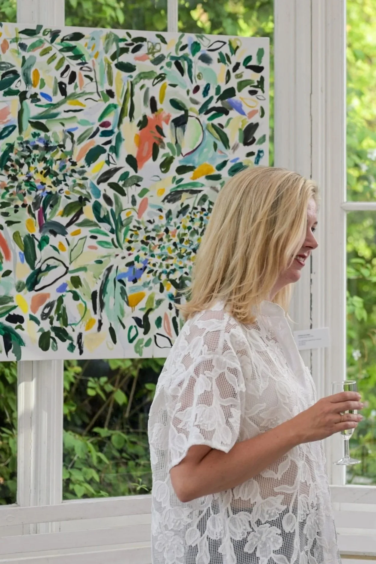 A woman with blonde hair wearing a white lace dress holding a glass of champagne, standing in front of a colorful abstract floral art piece on a bright day.