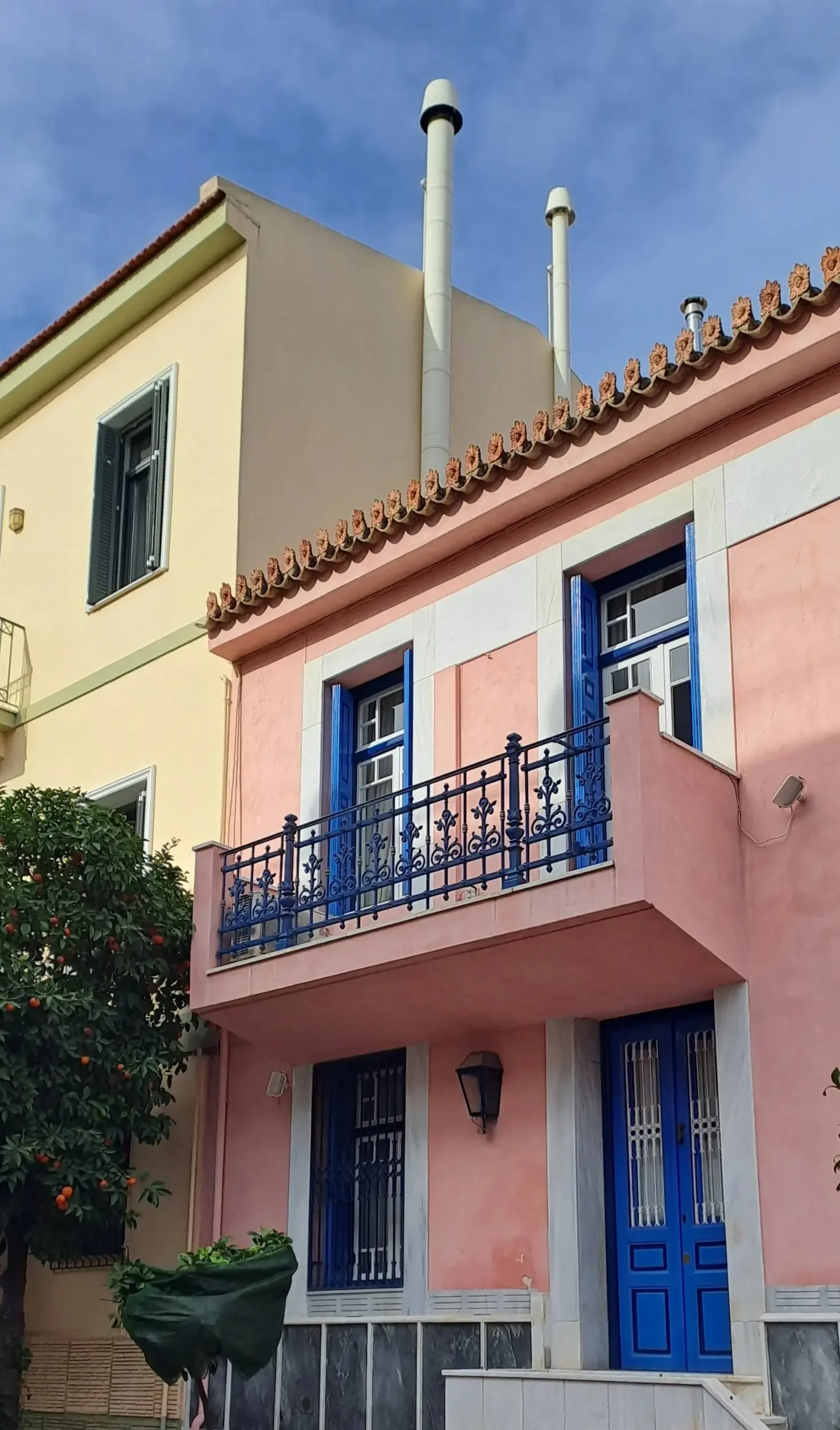 A colorful house with a pink exterior, blue door and window shutters, a small balcony with ornate black railing, and a balcony light fixture. There is a green tree with orange fruits to the left and a potted plant in the foreground. The sky is blue with a few clouds.