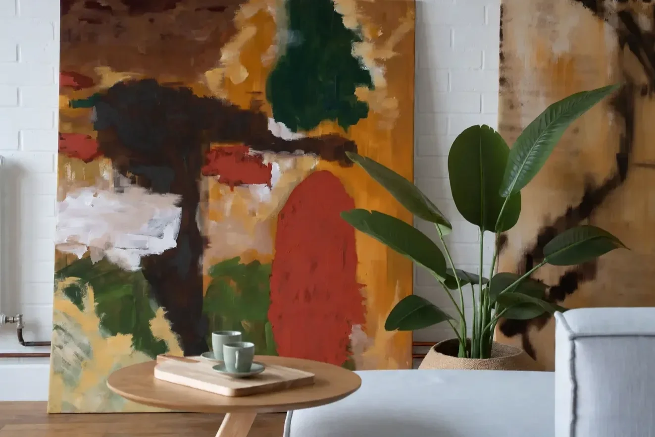 An abstract landscape painting with trees and a red hill, placed behind a potted plant with large green leaves, next to a small round table with three cups on it.