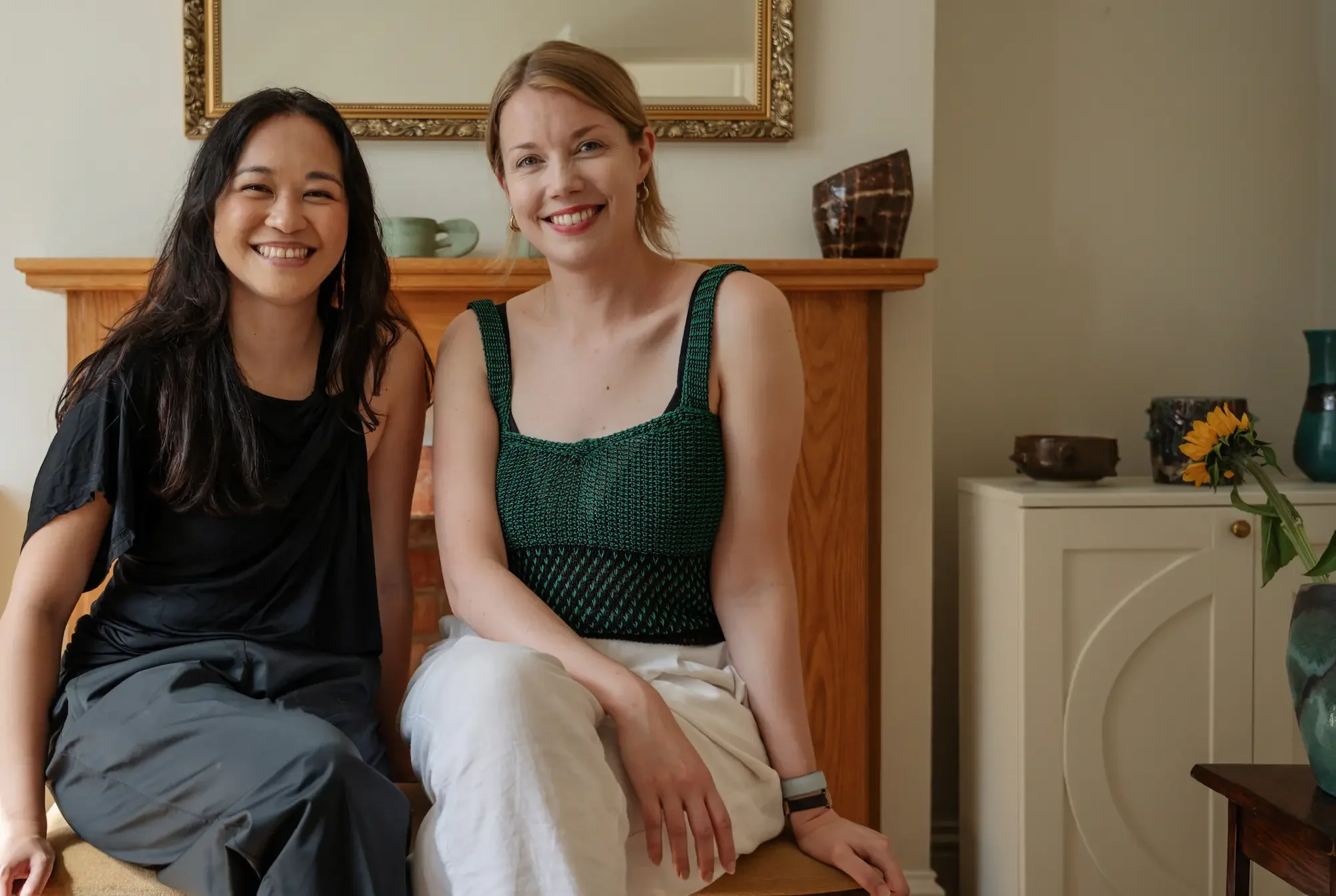 Two women sitting together in a cozy room, smiling at the camera. One woman has long dark hair and is wearing a black top, while the other has short light hair, and is dressed in a green and black patterned tank top and white pants. Behind them is a fireplace mantel with decorative items and a framed mirror on the wall.