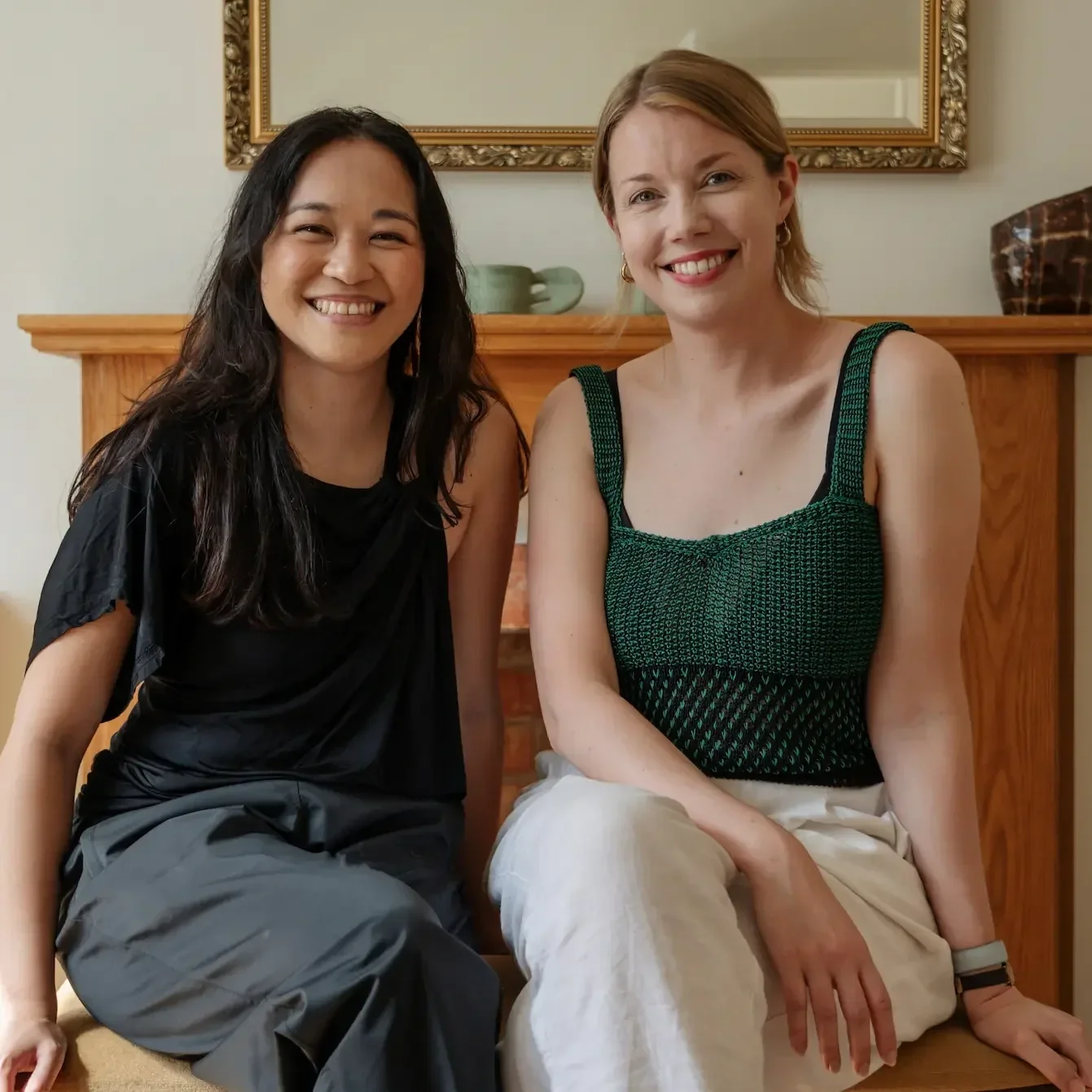 Two women sitting on a wooden bench in front of a mirror, smiling at the camera, in a warmly lit room with a wooden mantle and decorative items.