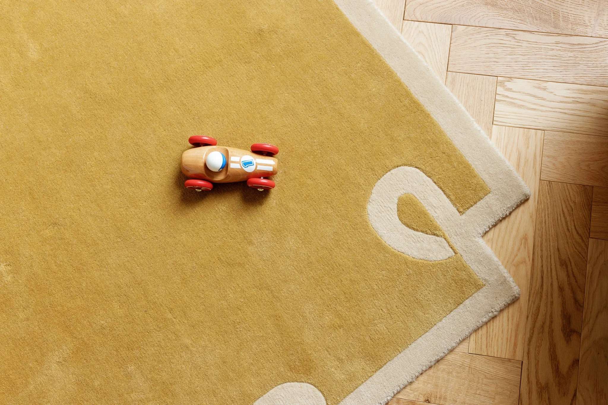 A small wooden toy car with red wheels and a blue and white logo on its side, positioned on a yellow and white patterned rug on a wooden floor.