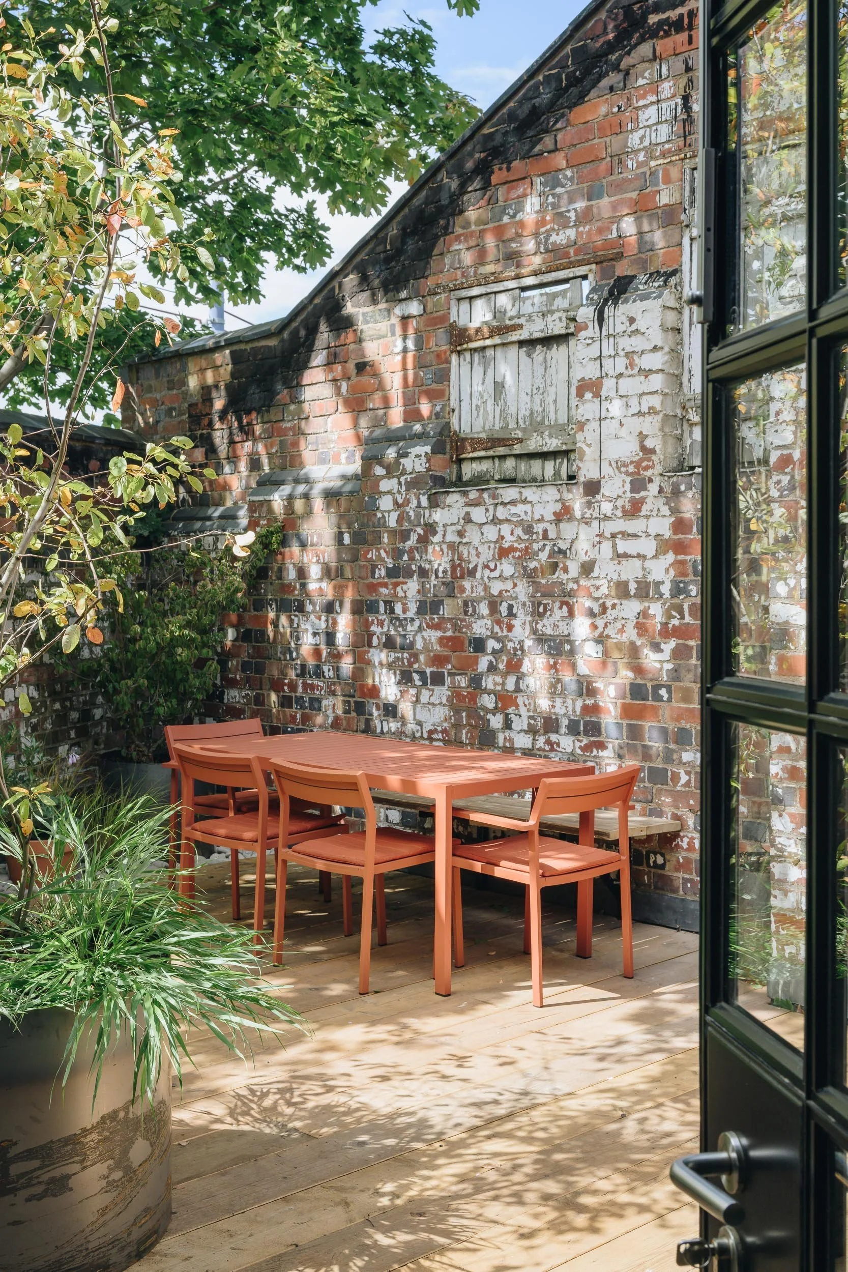 View of a cozy outdoor patio with a wooden dining table and six matching chairs, positioned on a wooden deck, next to an old brick wall with a window boarded up with white-painted wood. The patio is surrounded by lush green plants and trees, with sunlight creating patterned shadows on the deck.