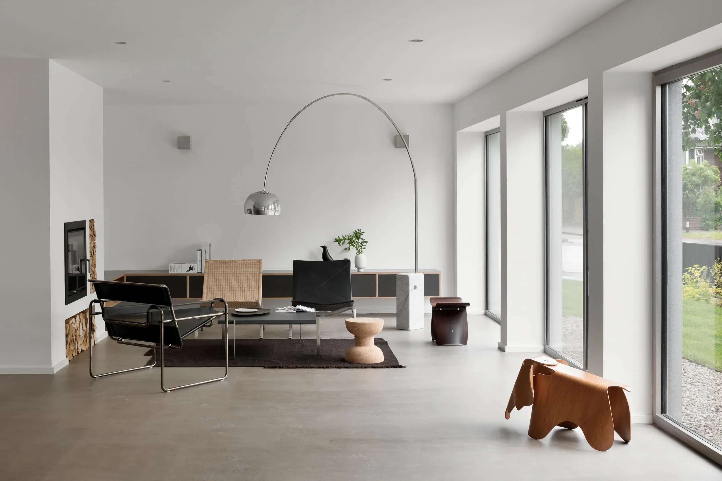 Modern living room with large windows, minimalist furniture, a black armchair, wooden chairs, a small plant, a white decorative vase, and an arched floor lamp with a metallic shade.