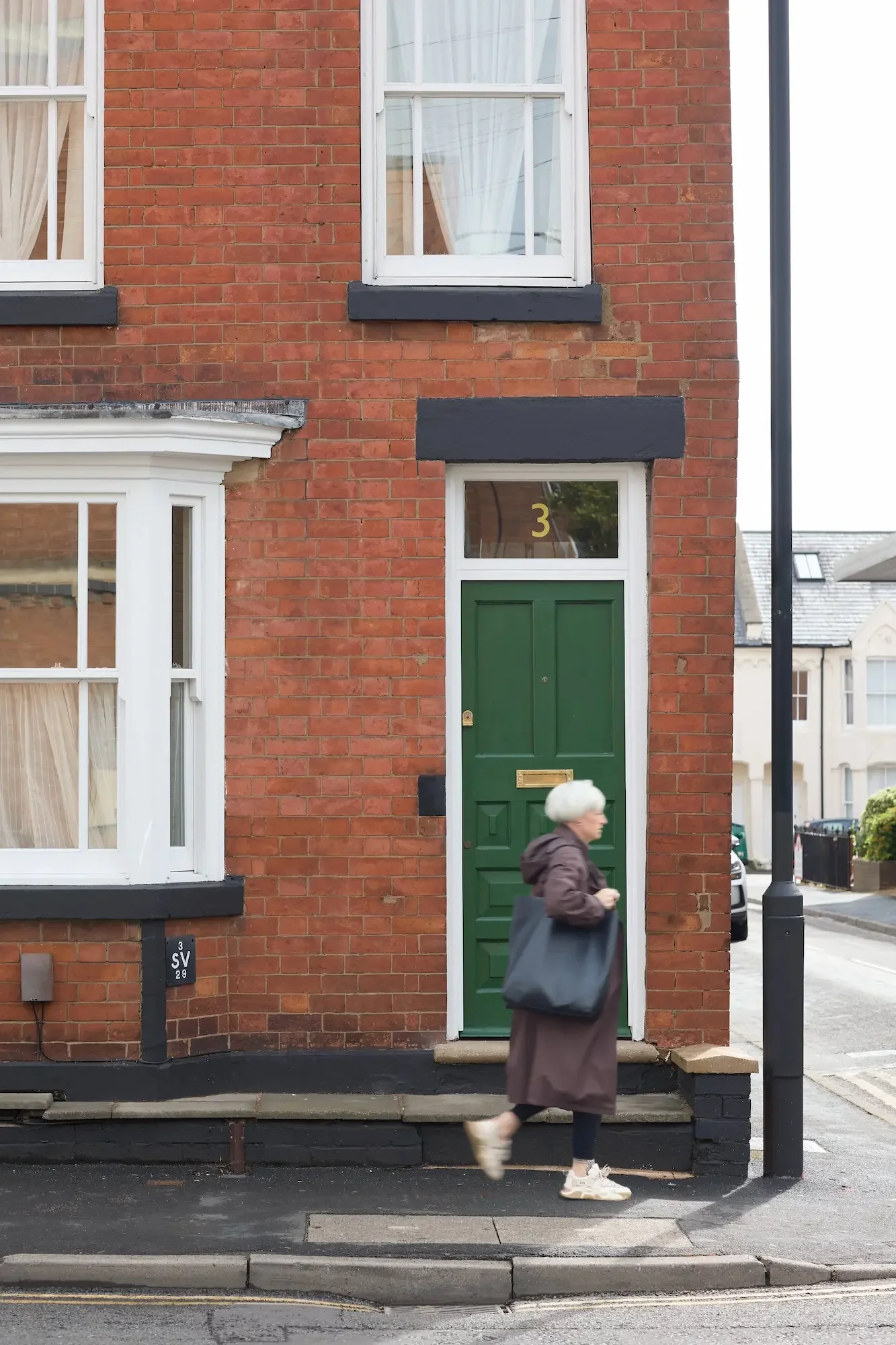 A brick building with black accents and a green front door numbered 3. A person with white hair, wearing a long brown coat and white sneakers, is walking past the building on the sidewalk.