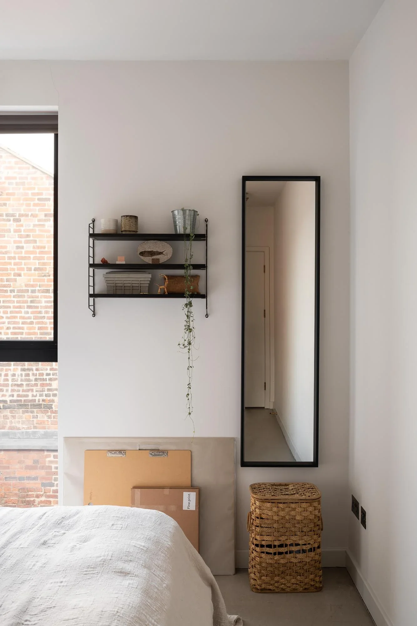 A bedroom with a white bed, a black wall-mounted shelf with decorative items, a tall rectangular mirror, a woven basket, and a window showing a brick building outside.