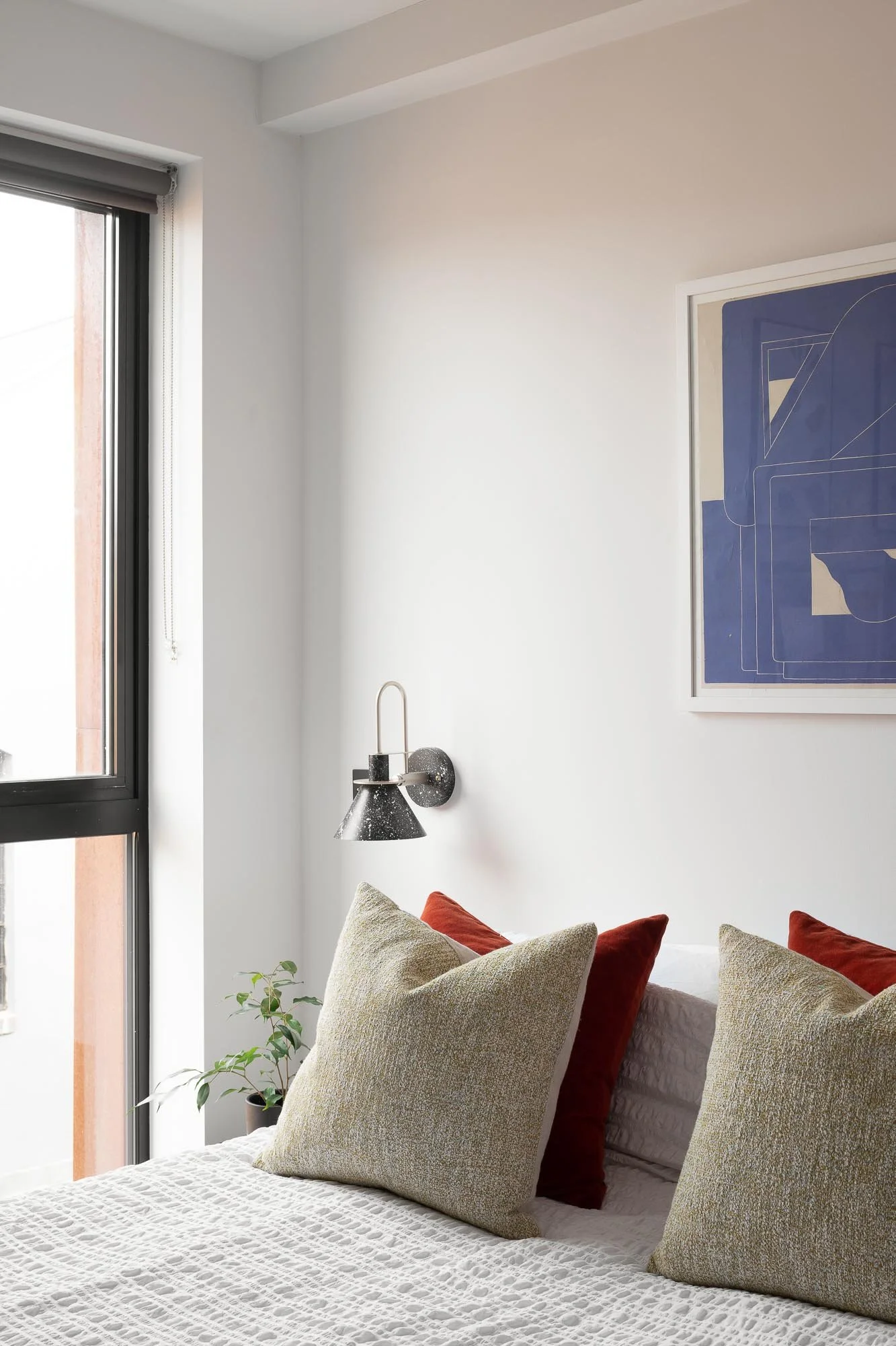 A bedroom with a large window, a white bedspread, and pillows in beige and rust colors. A black wall-mounted lamp is beside the bed, and a blue geometric artwork hangs on the white wall. A small green plant is near the window.