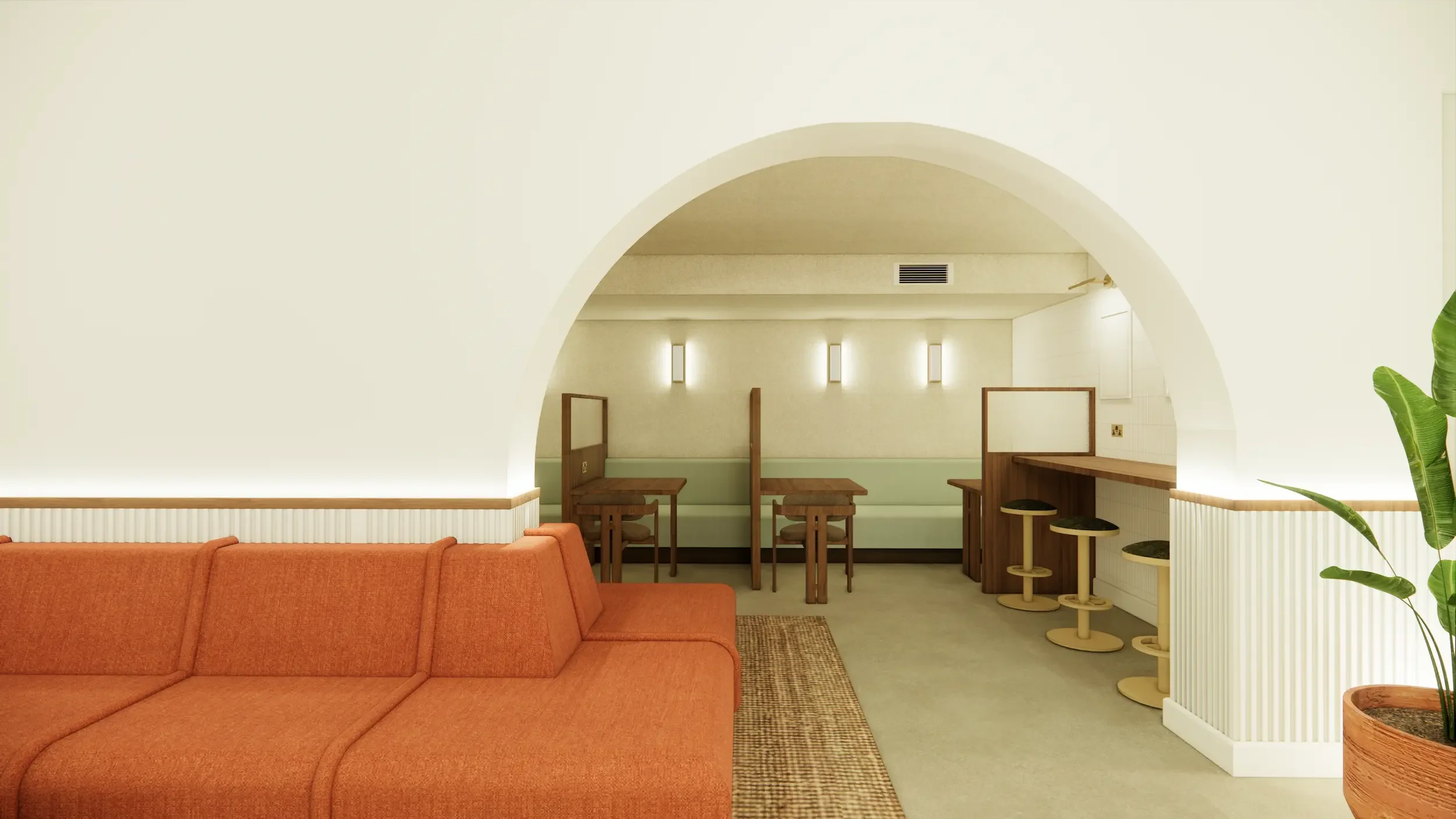 A cozy indoor space with a burnt orange sofa in the foreground and a small dining area with wooden chairs and a bar seating area with stools in the background. The room has cream-colored walls, soft lighting, and a large green plant on the right.